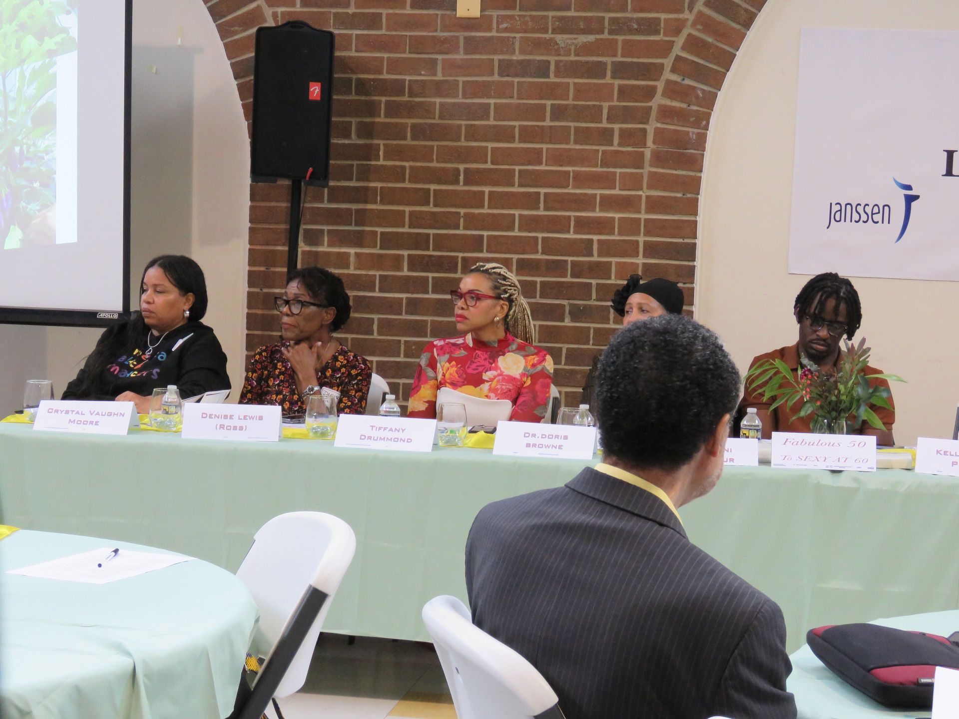 Panel discussion at a table with name placards. Speakers and audience visible in a room with a brick wall.