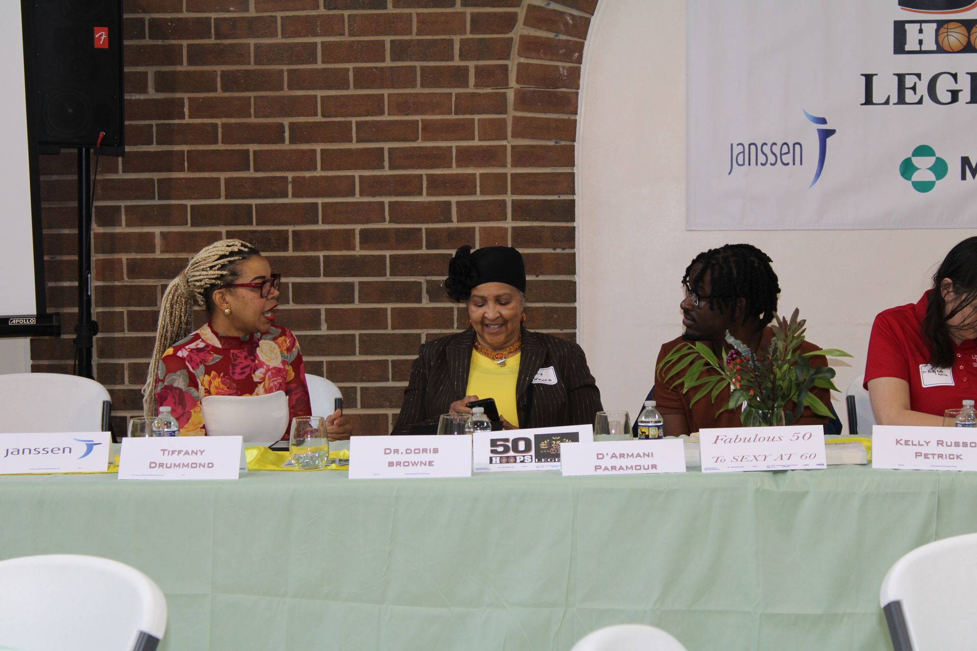 Panel discussion at a table with brick backdrop. Four people sit, speaking. White banner with sponsor logos.