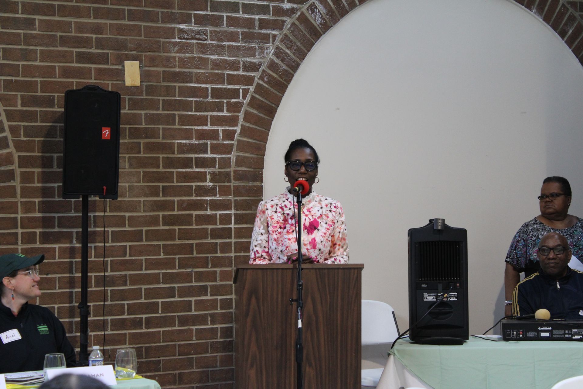 Woman speaking at a podium with microphone in front of brick wall. Other people sit at a table.