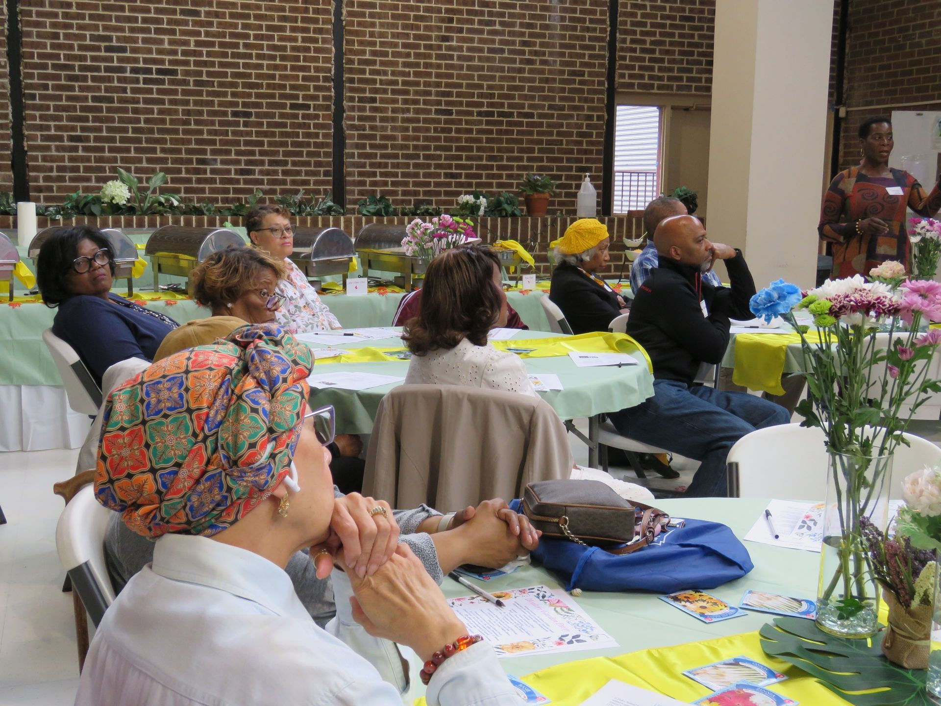 People seated at tables, listening to a speaker in a room with brick walls. Flowers on tables.