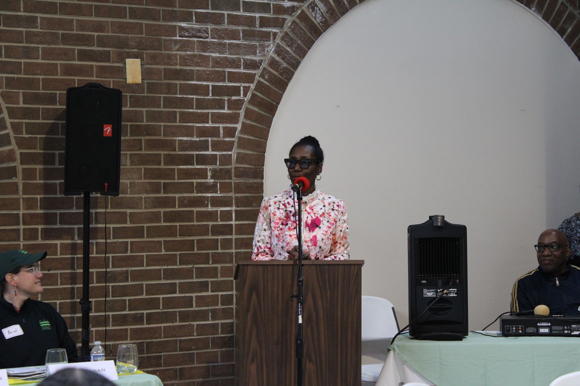 Woman speaking at a podium with two speakers and people seated at a table in a brick-walled room.