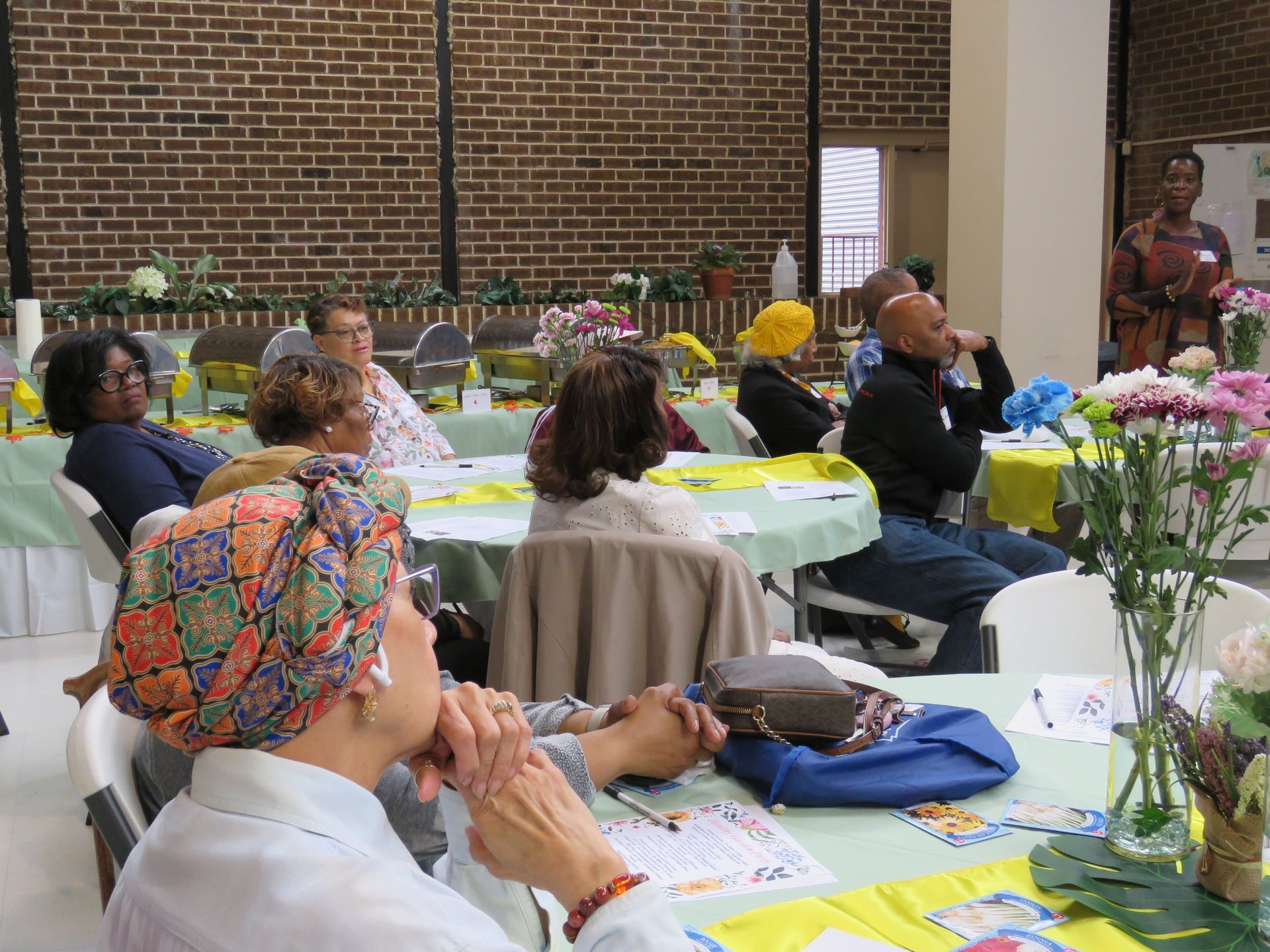People seated at tables, listening to a speaker in a room with a brick wall. Floral arrangements and food present.