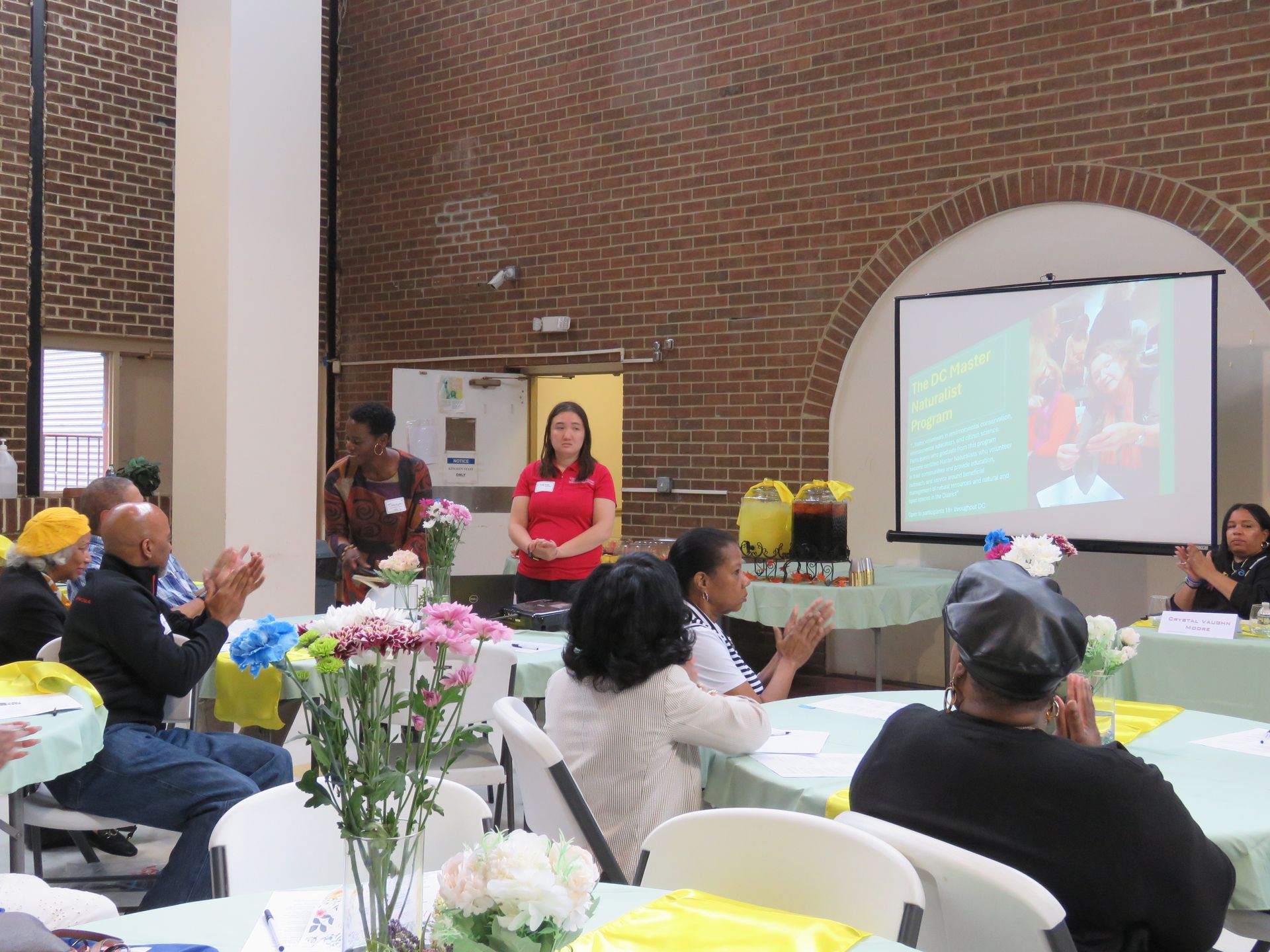People seated at tables, applauding a woman speaking near a screen, in a brick-walled hall.