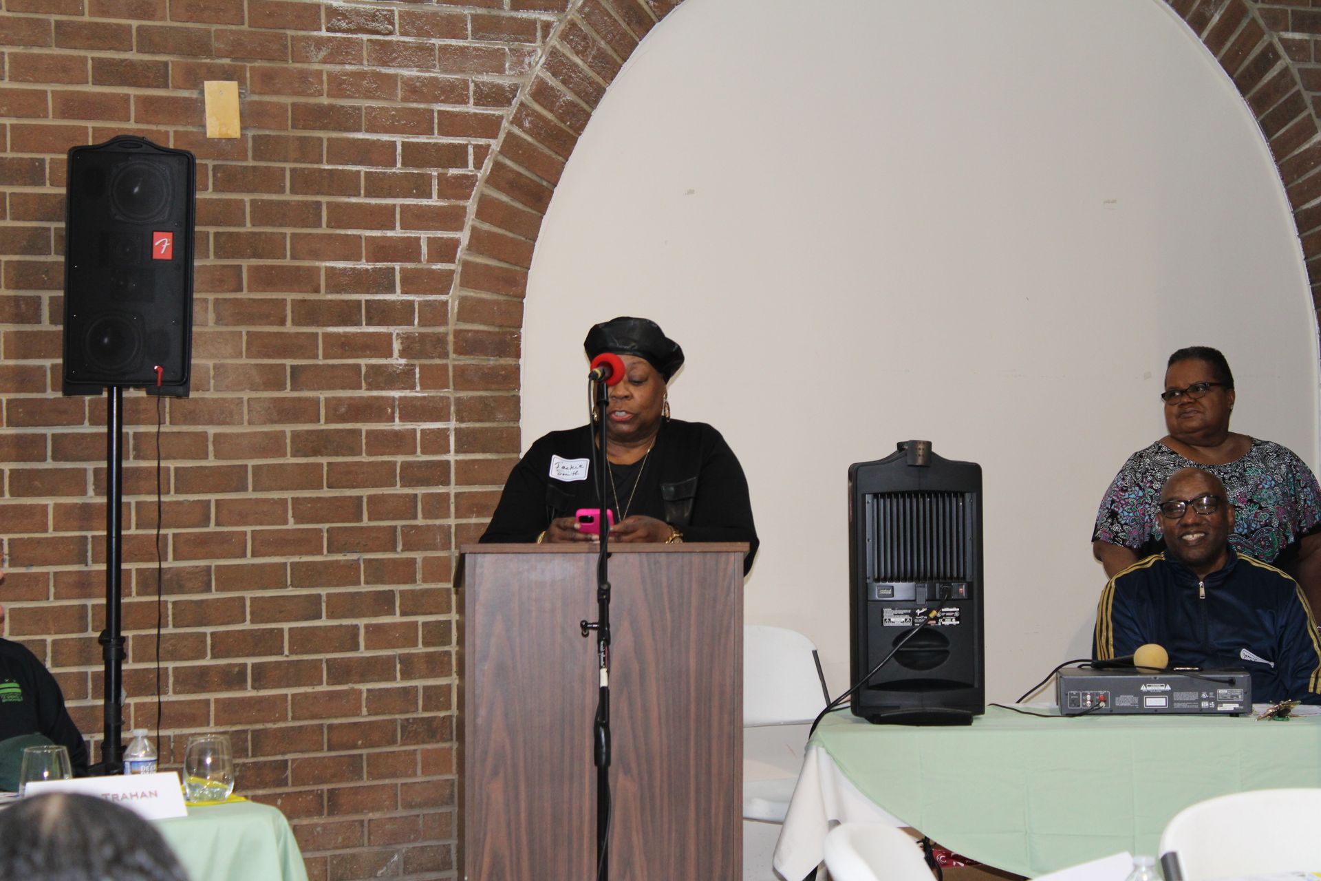 Woman speaking at podium during event; brick wall and arched doorway in background.