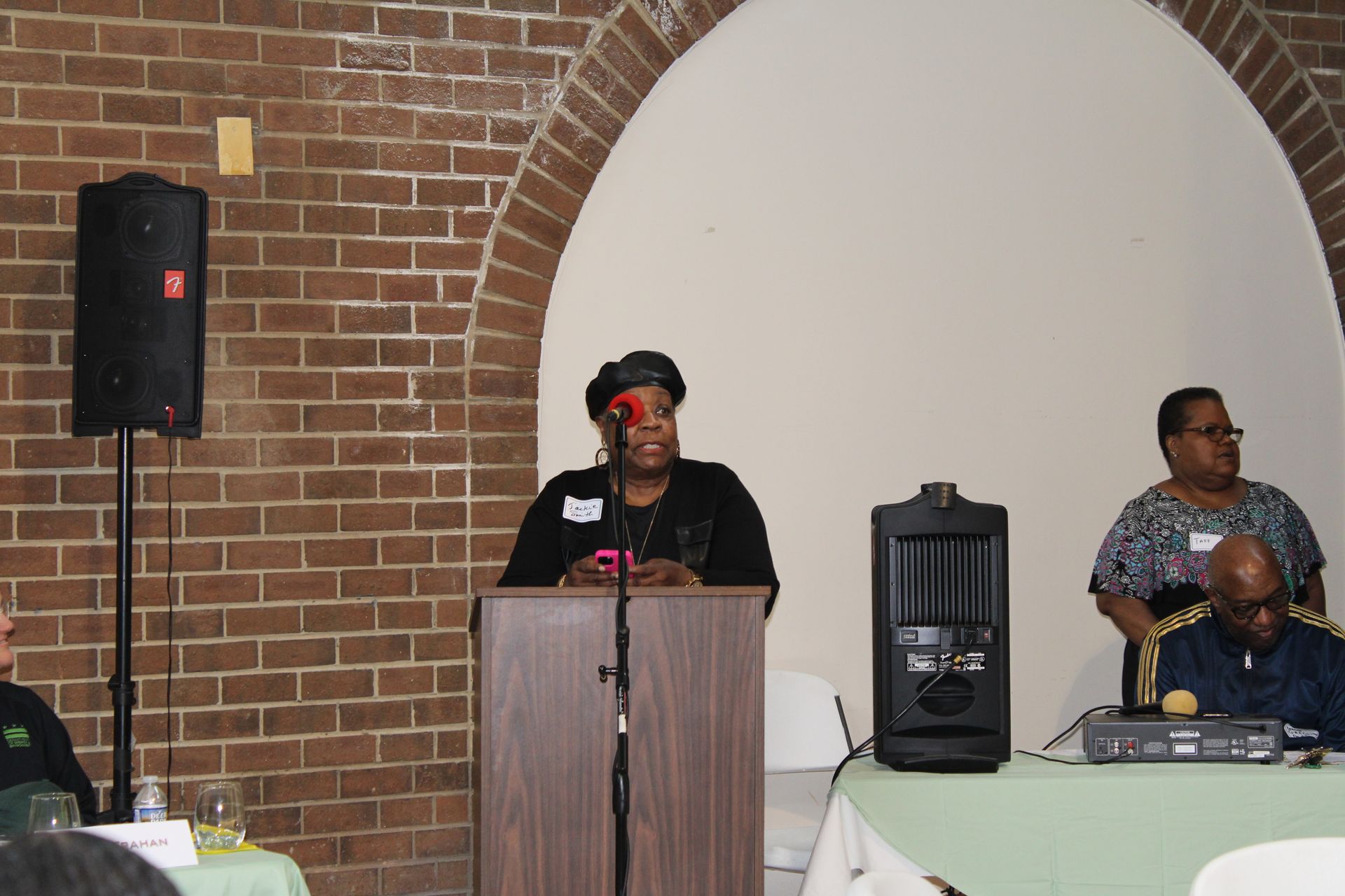 Woman speaking at a podium, brick wall background, another speaker to the right.