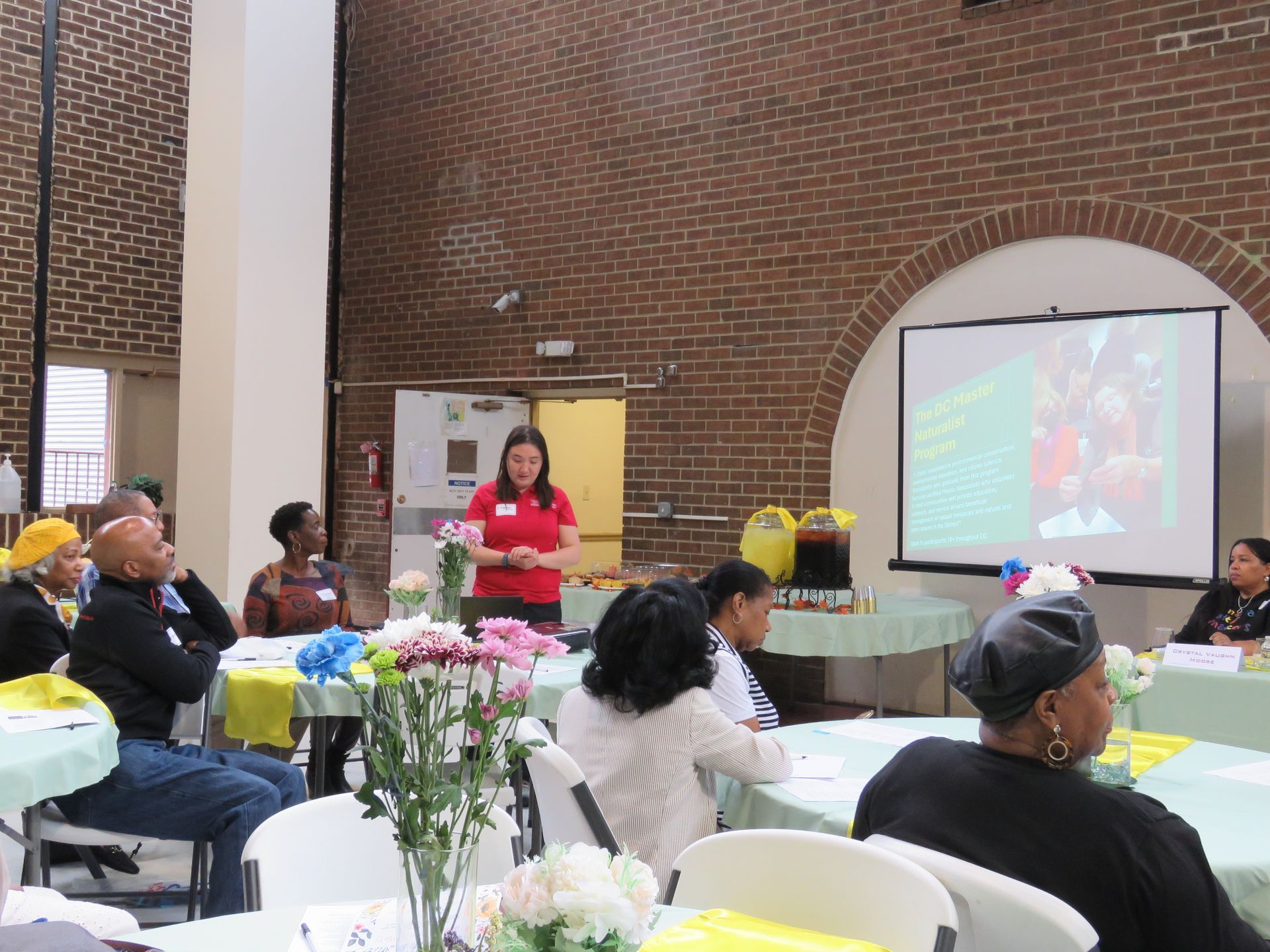 Woman presenting at a meeting with attendees seated at tables in a room with a brick wall.