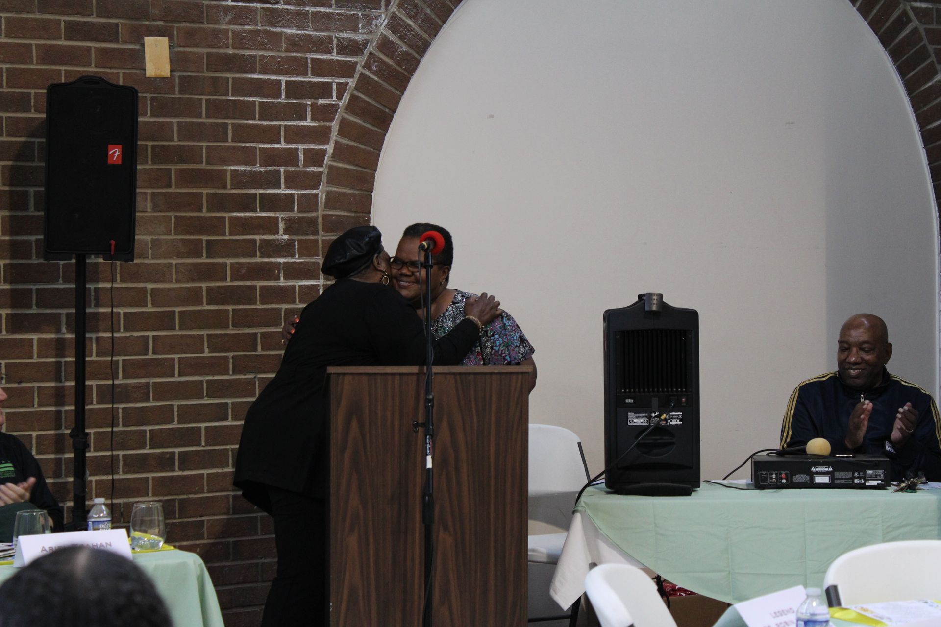 Two women embracing at a podium, brick wall background, another person at a table, possibly at an event.