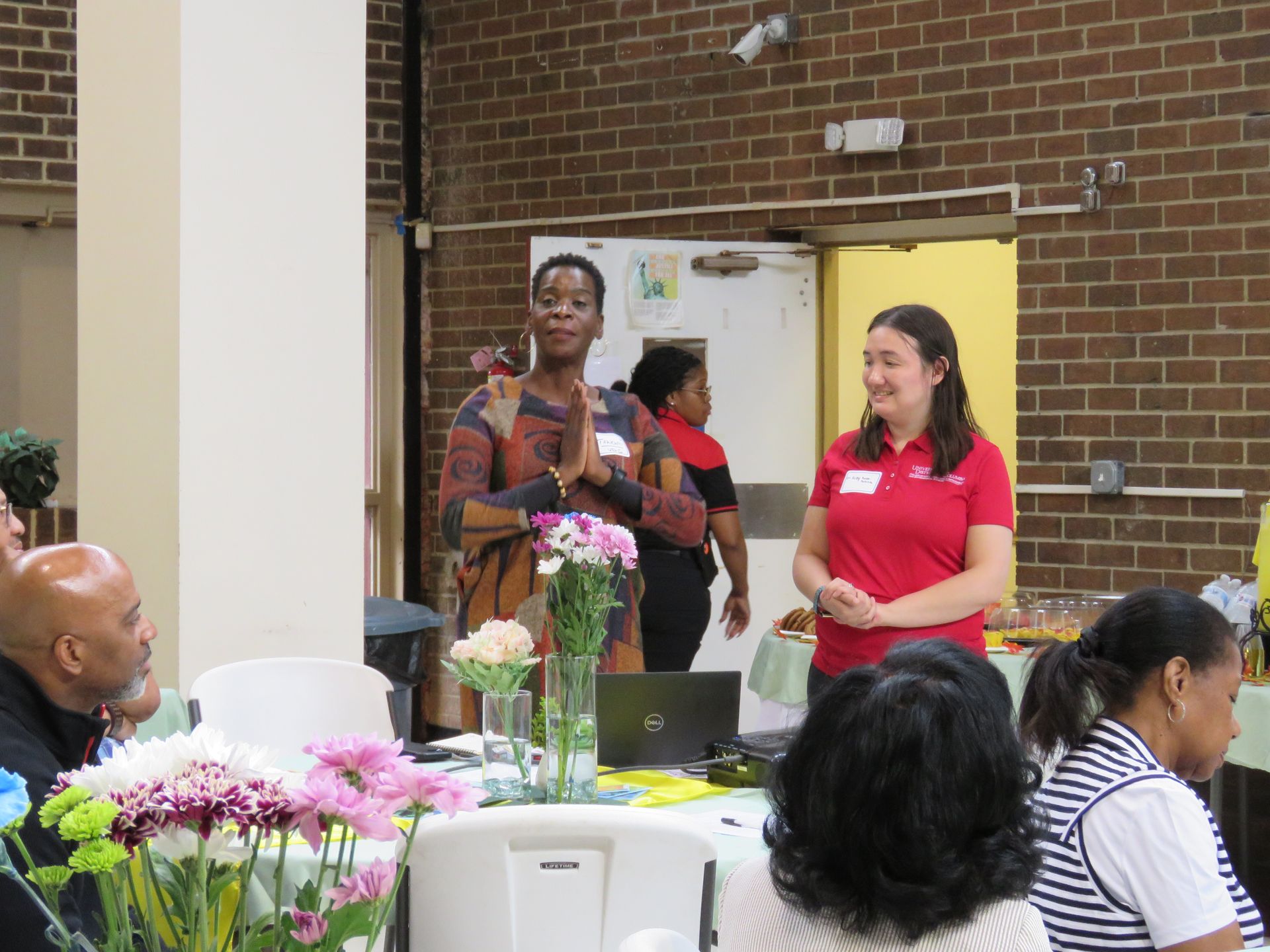 Two women at a presentation, flowers on table, people seated in a brick-walled room.