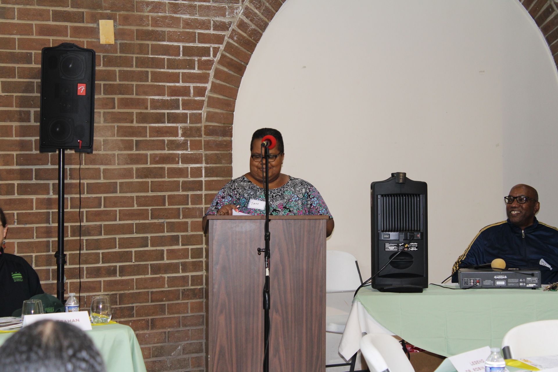 Woman speaking at a podium in front of a brick wall, speaker and audience members seated at tables.
