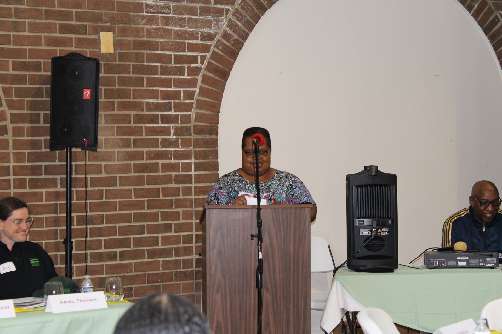 Woman speaking at a podium, flanked by two people at a table, brick wall background.