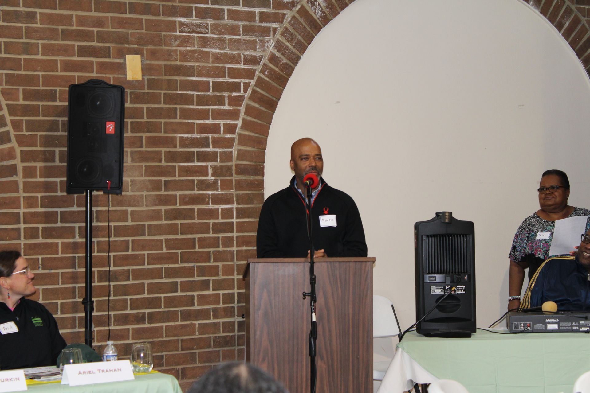 Man speaking at a podium with two people seated nearby. Brick wall and arched alcove in background.