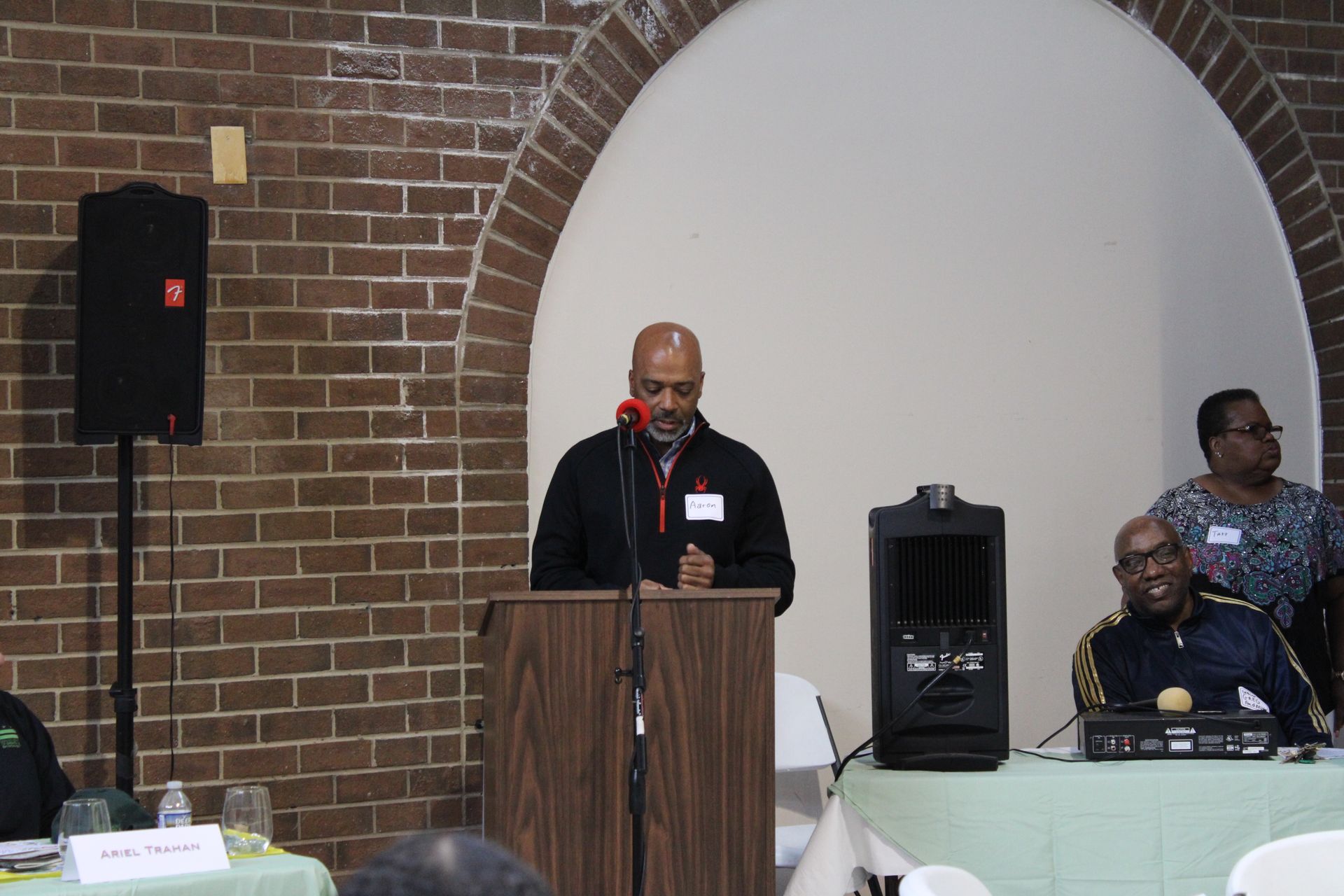 Man speaking at a podium in a brick-walled room; another man sits at a table to the right.
