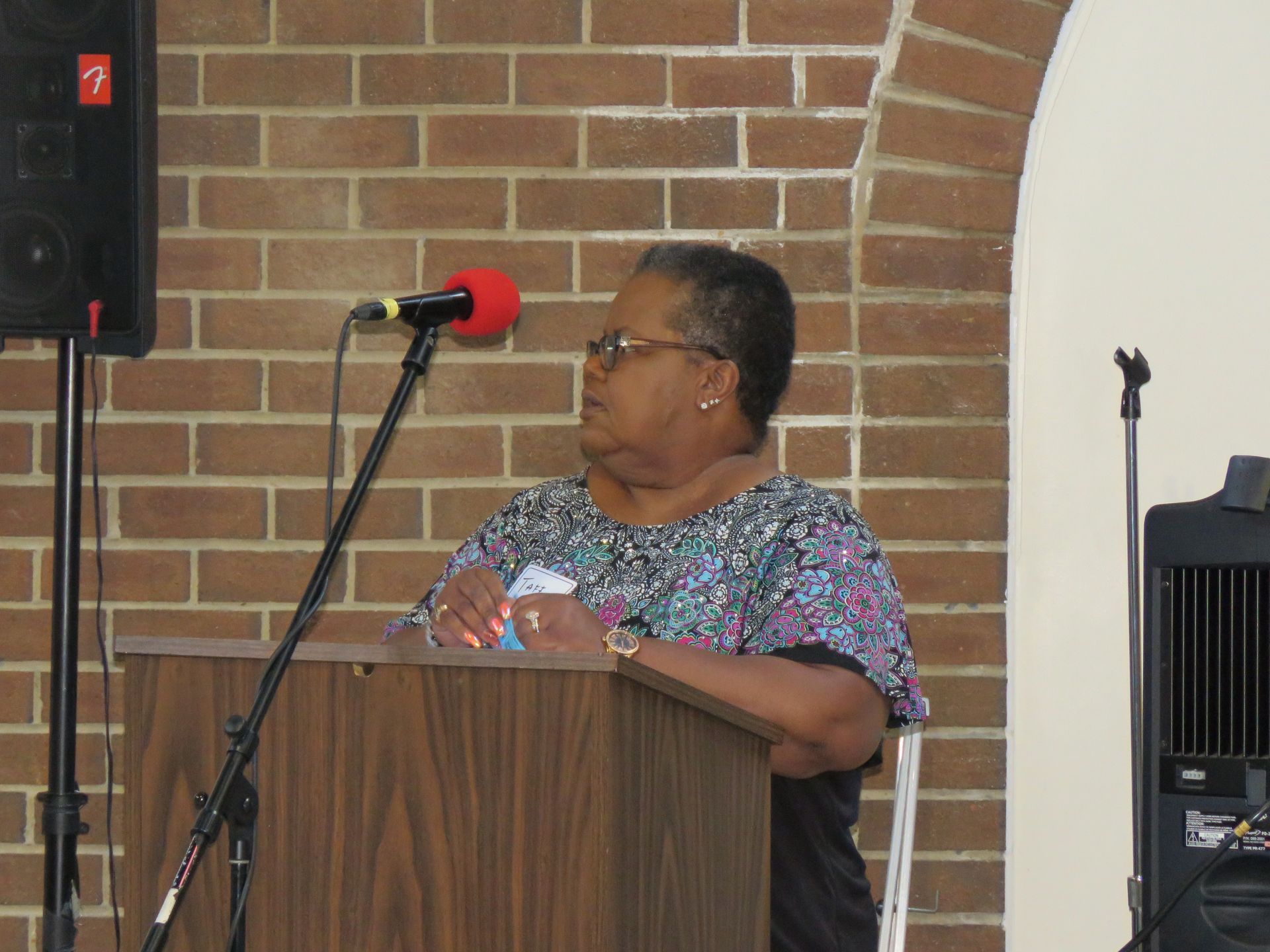 Woman speaking at a podium in front of a brick wall and speaker.