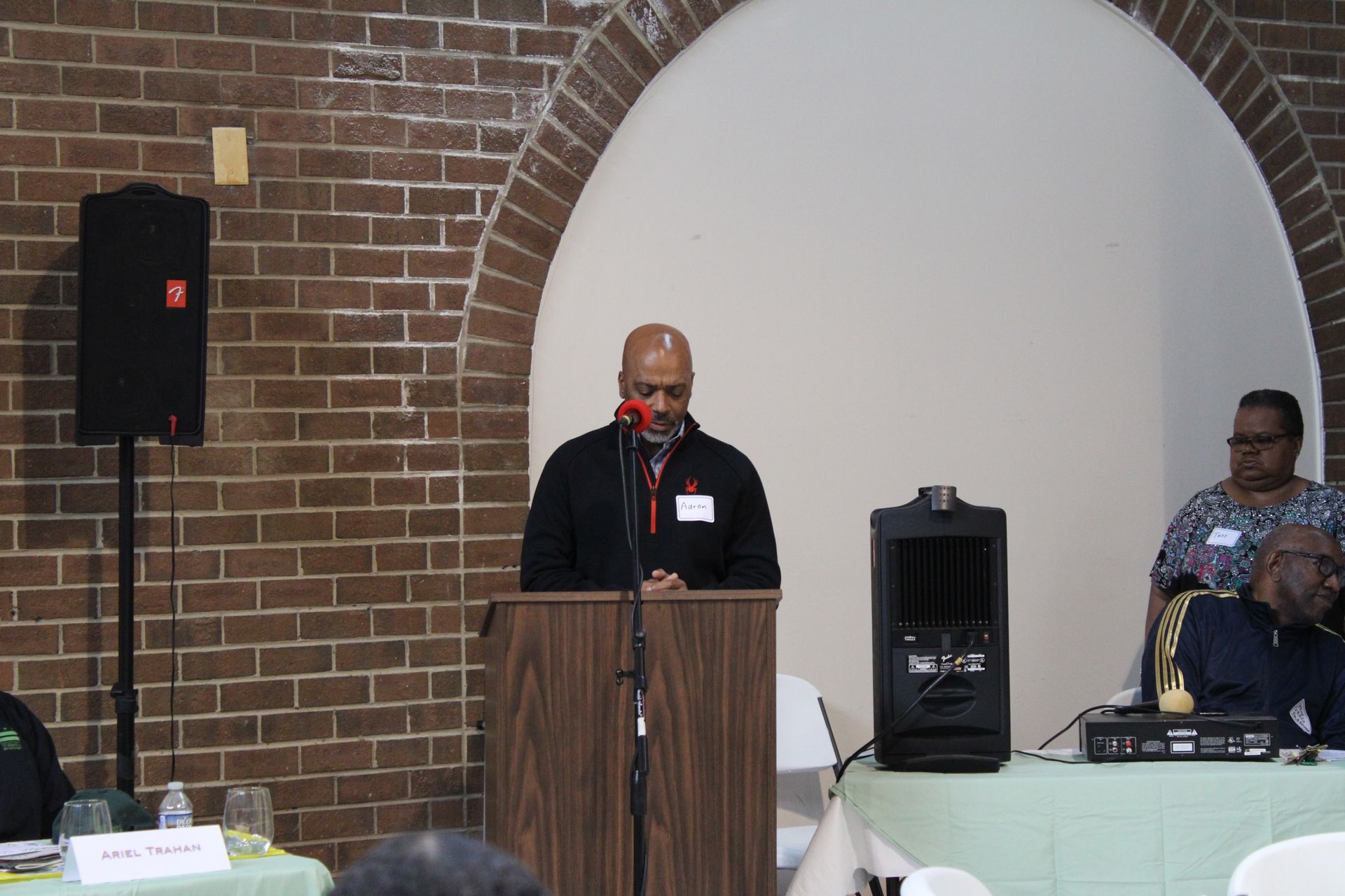 Man speaking at a podium in a room with a brick wall and seated audience.