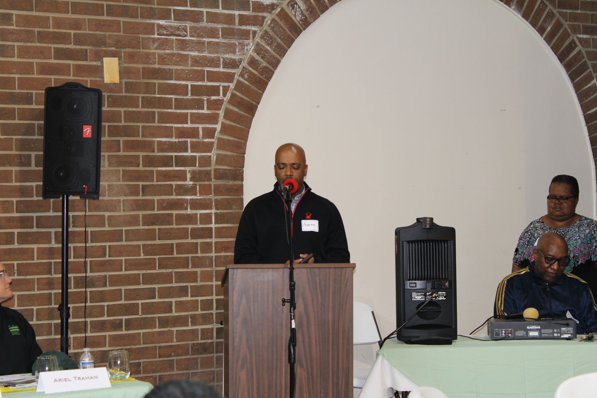 Man speaking at a podium with microphone in front of a brick wall and arched doorway. Two people seated behind.