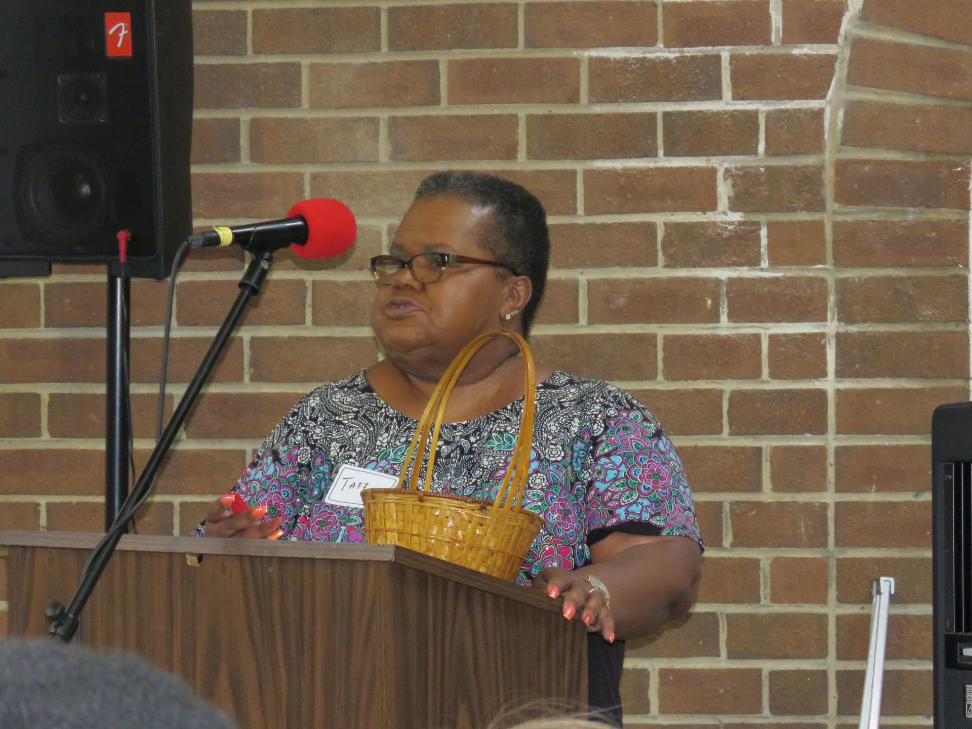 Woman speaking at a podium with microphone, holding a basket, in front of a brick wall.