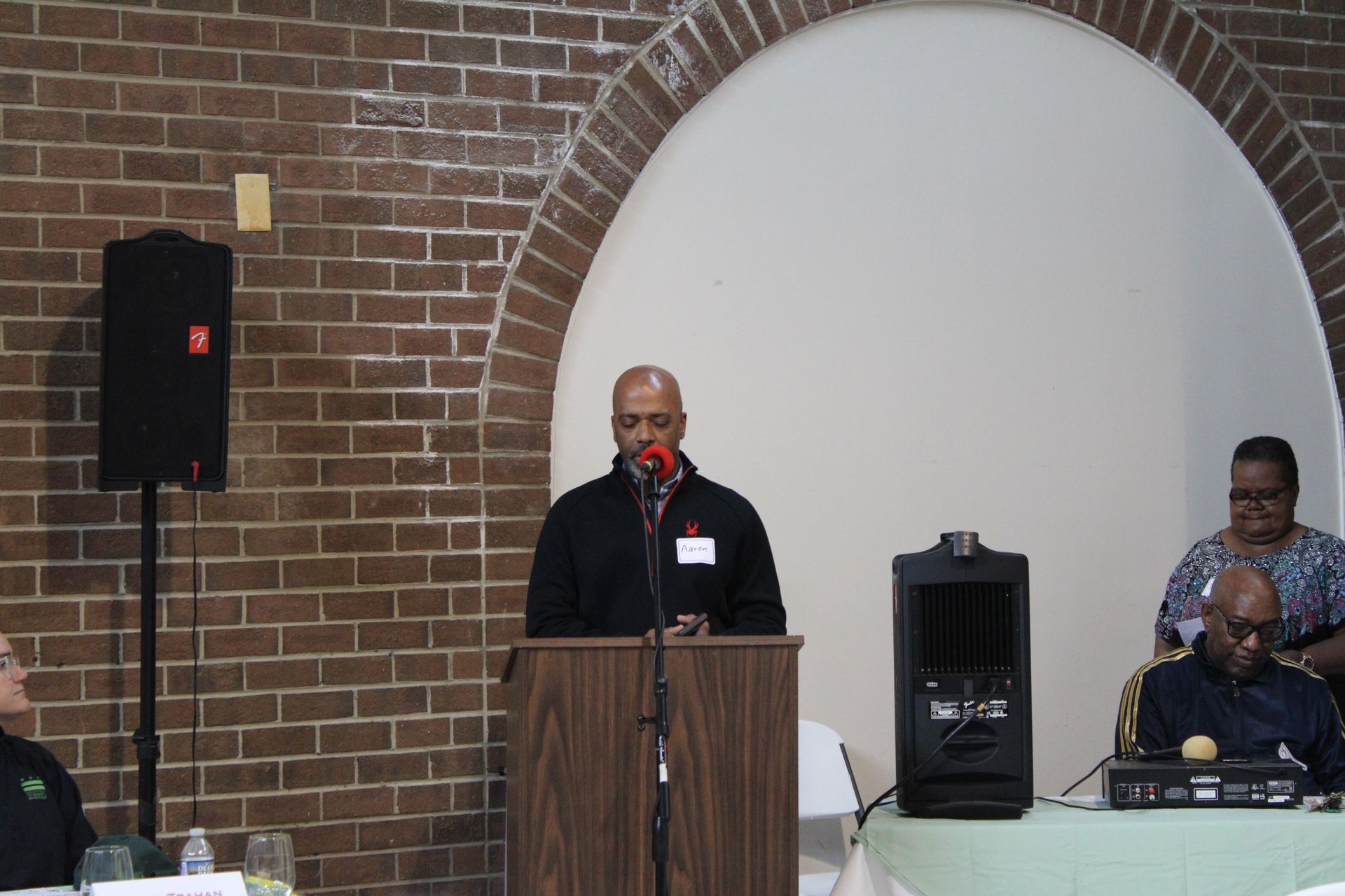 Man speaking at a podium, brick wall backdrop. Speakers and other people are visible nearby.