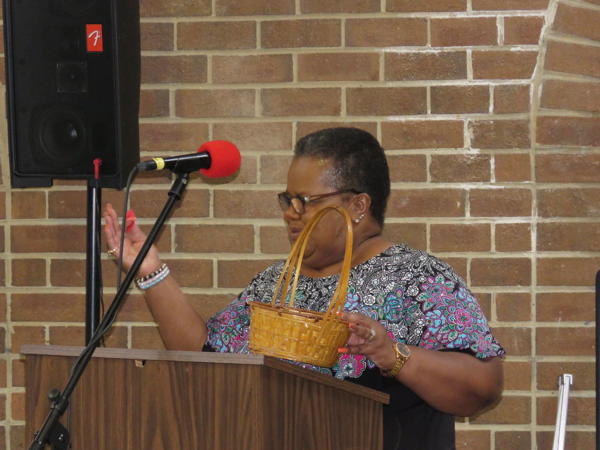 Woman at a podium with a basket, speaking near a microphone in front of a brick wall.