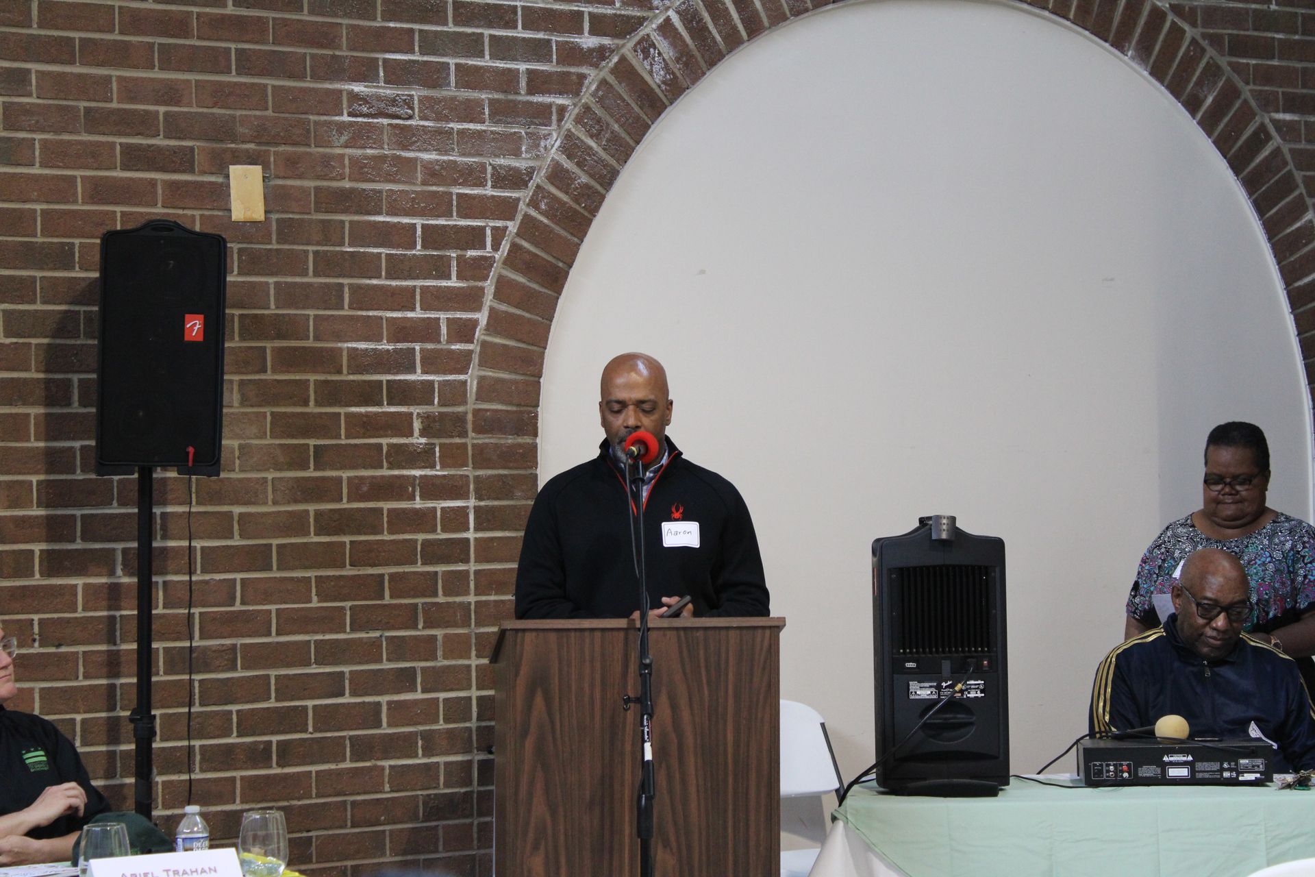 A man speaking at a podium in front of a brick wall and seated audience.