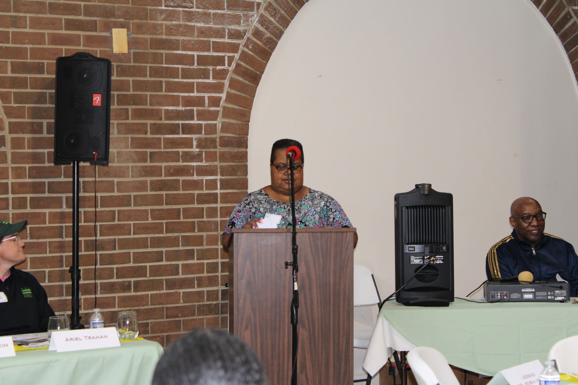 Woman speaking at a podium, two others at a table with green cloth, brick wall backdrop.