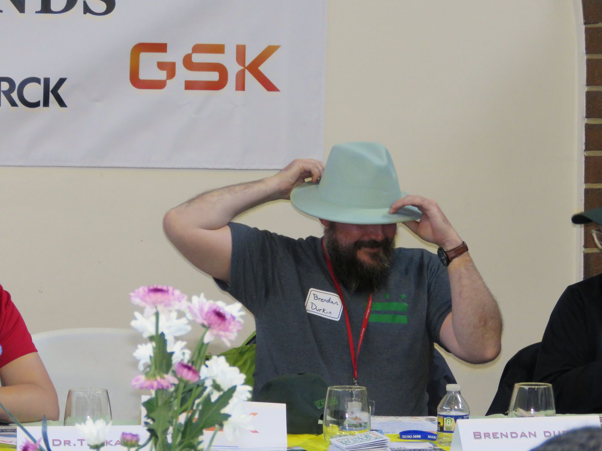 Man adjusting a light green hat, seated at a table with a banner reading 