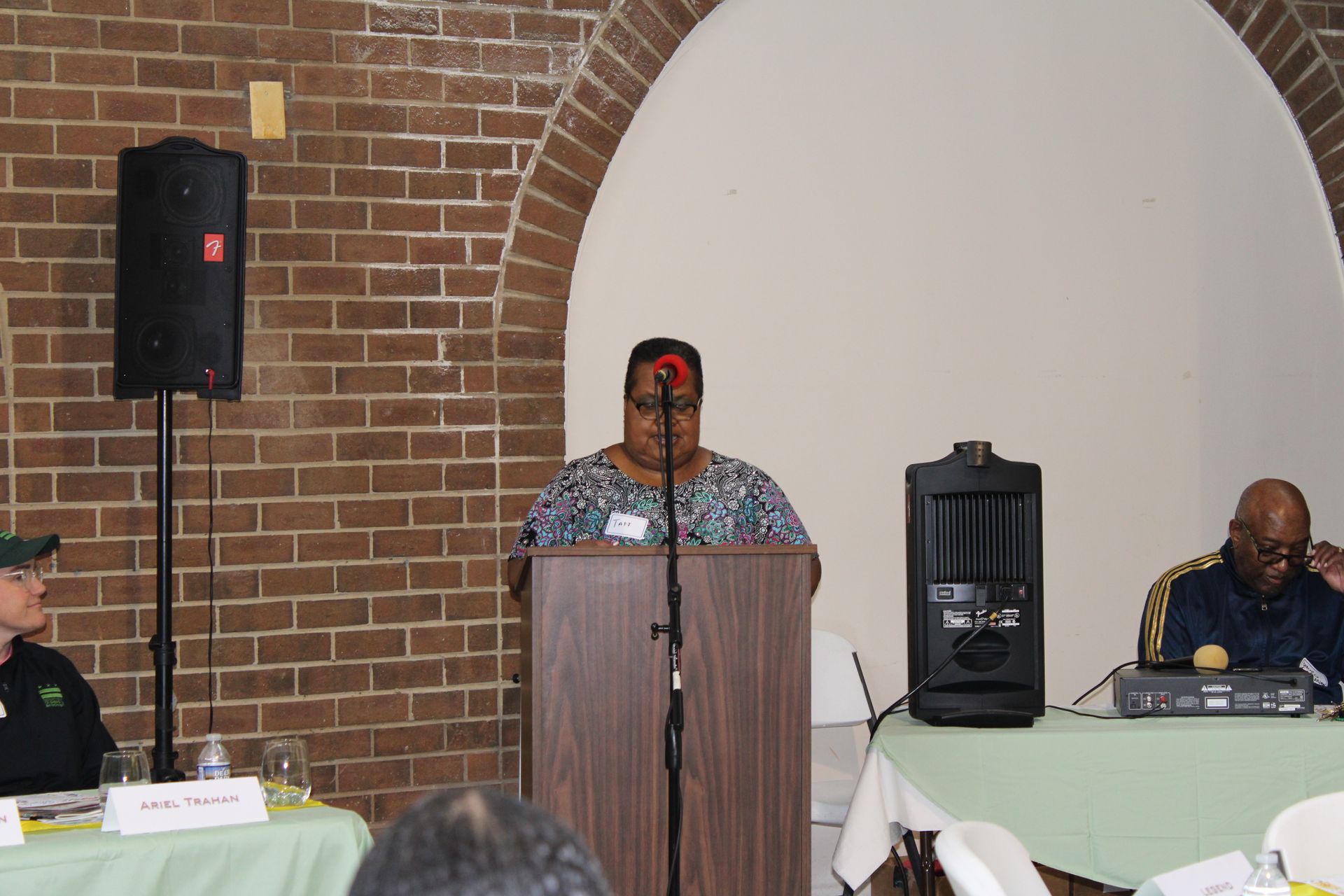 Woman speaking at a podium, flanked by two people seated at a table. Brick wall background.