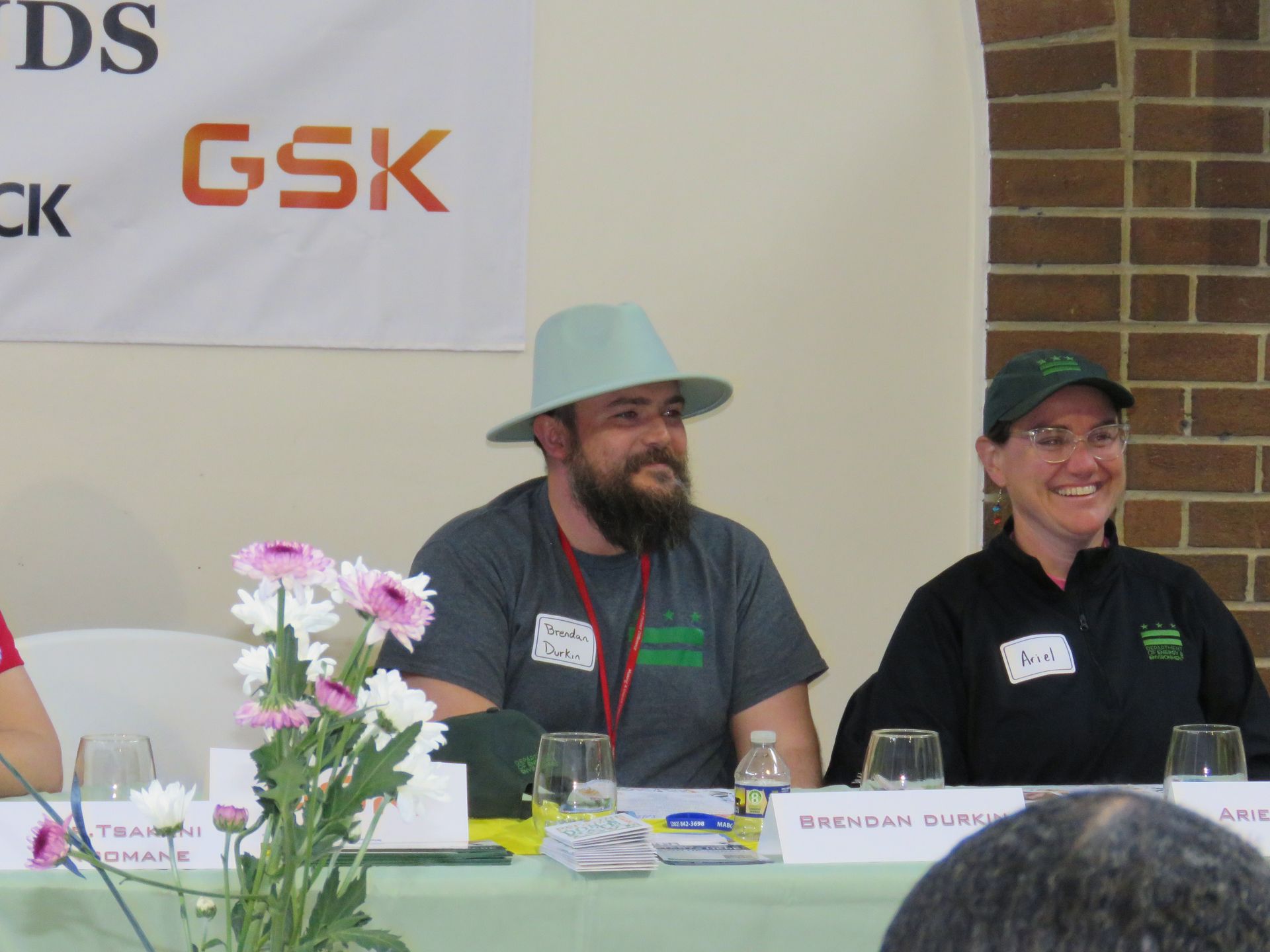 Two people seated at a panel, the man wearing a hat, the woman a cap. Both smiling. A banner with text in the background.