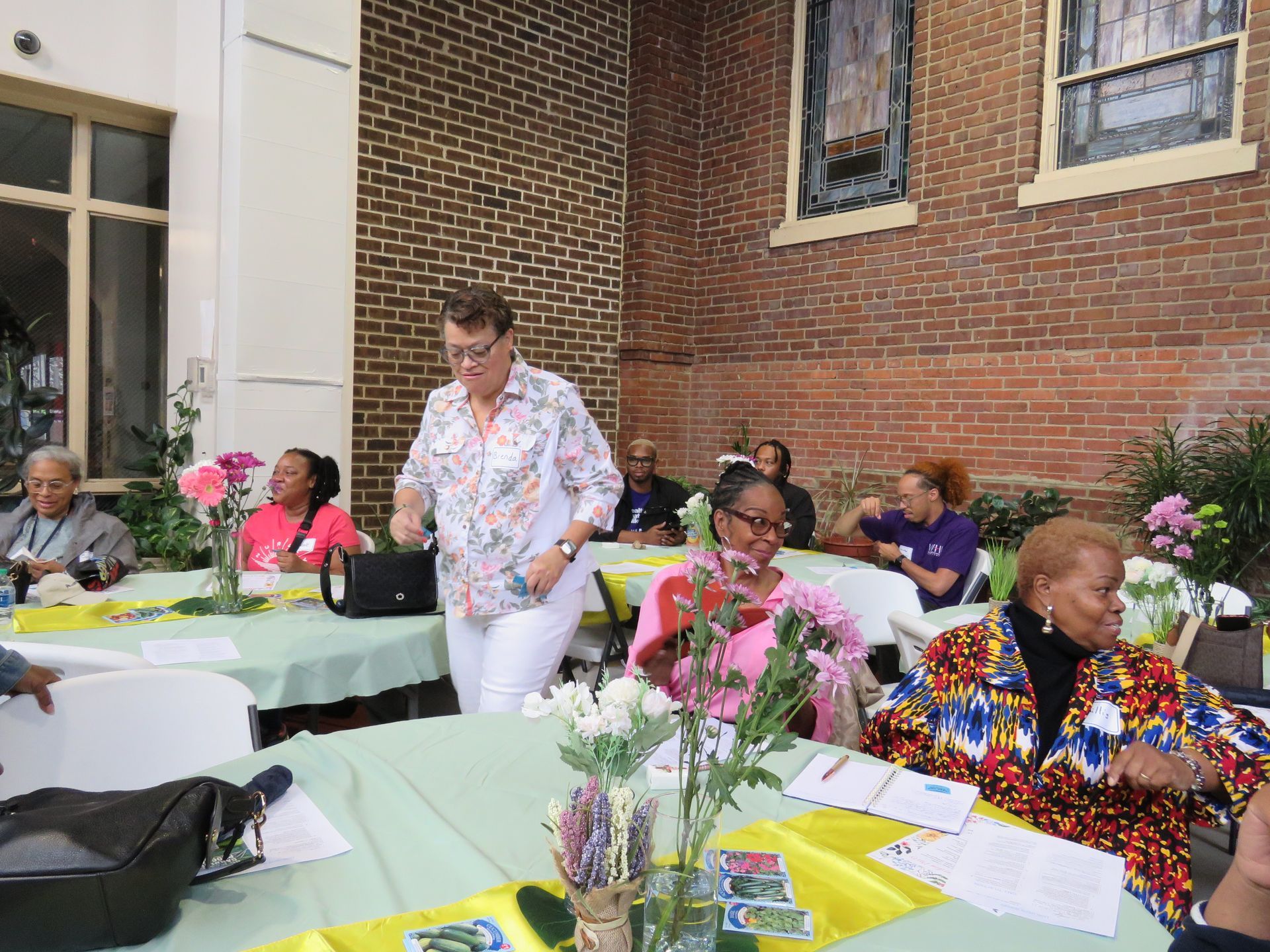People seated at tables in a courtyard, a woman walks by. Tables have green cloths, flowers. Brick wall in the background.