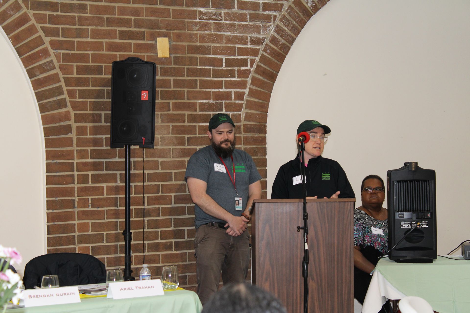 Two people speak at a podium in front of a brick wall, speaker and audience visible.