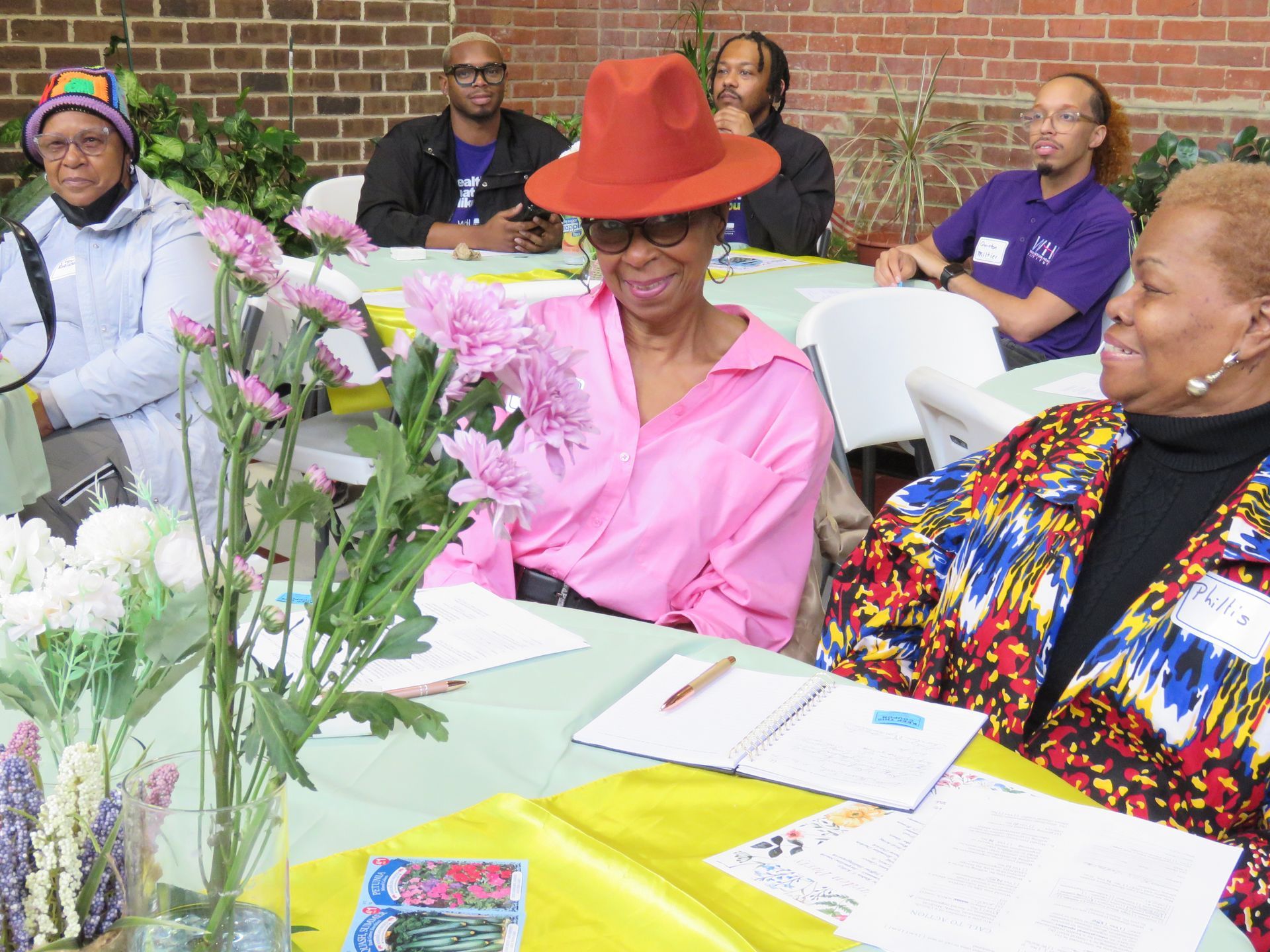 Group of people seated at a table with flowers. Woman in a pink shirt and hat interacts with others.