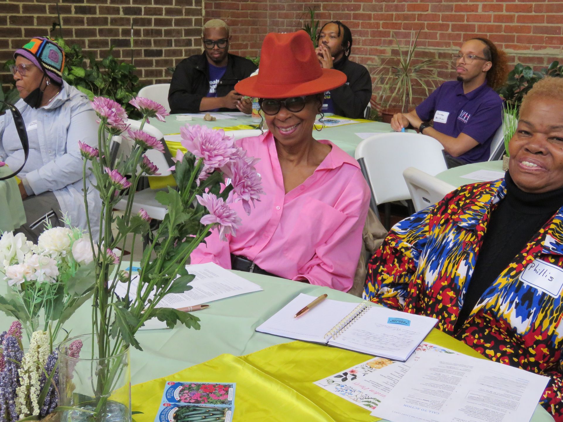 People seated at tables outdoors, smiling, with flowers, and writing materials.