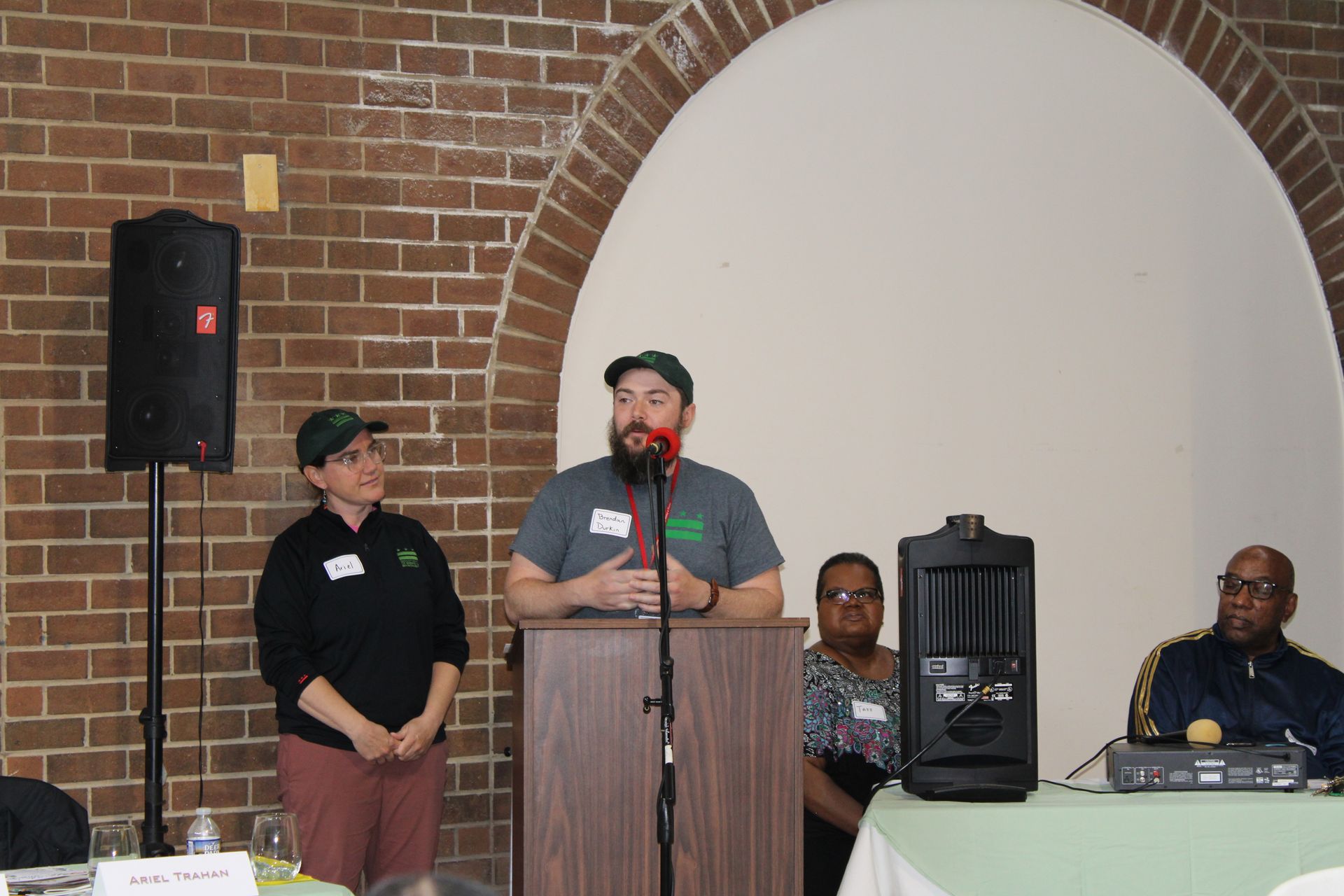 Man speaking at podium, two people beside and behind him, brick wall backdrop.