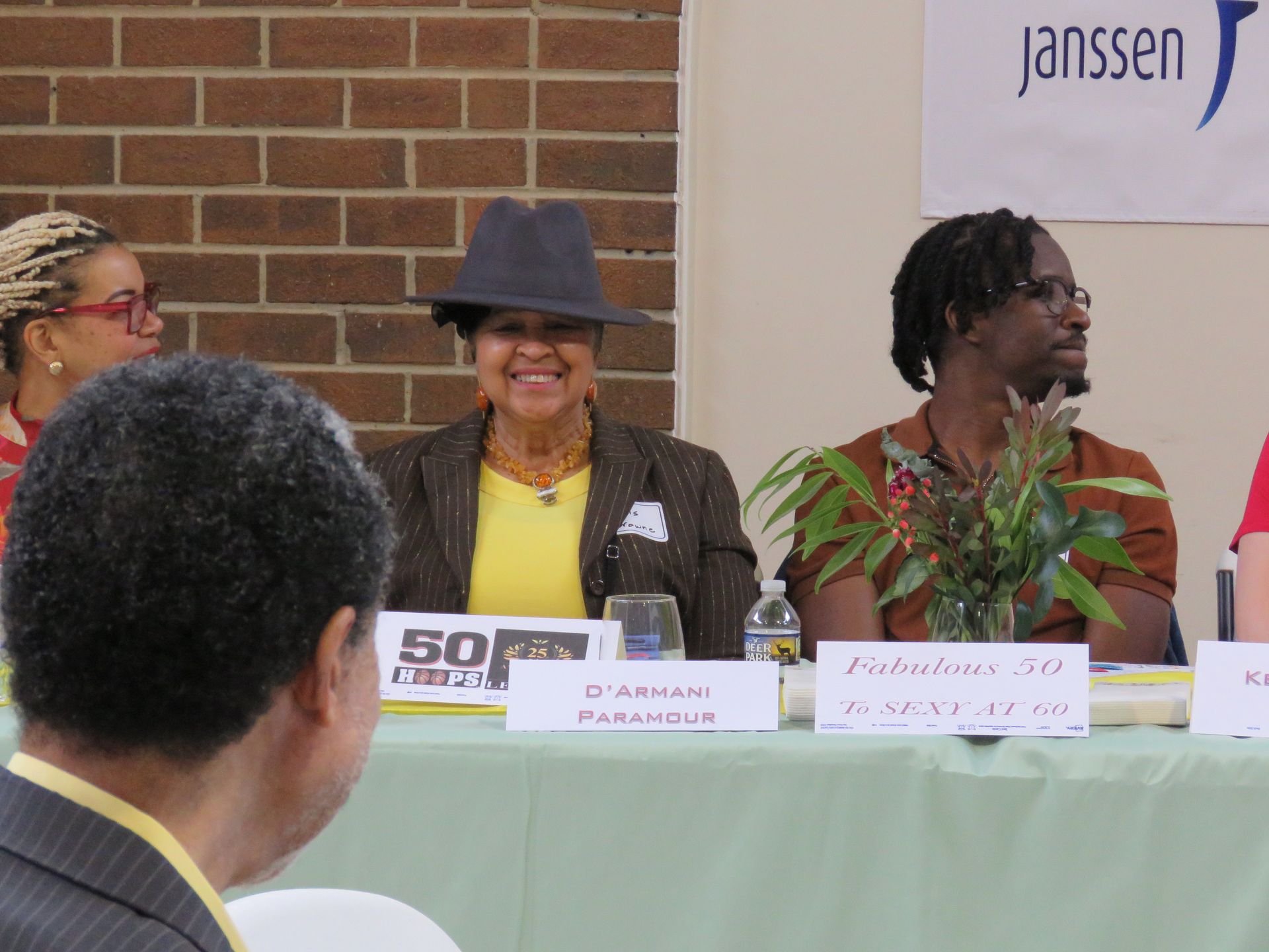 Panel discussion with seated speakers, brick wall backdrop. Speakers smiling and engaged, yellow and brown tones.