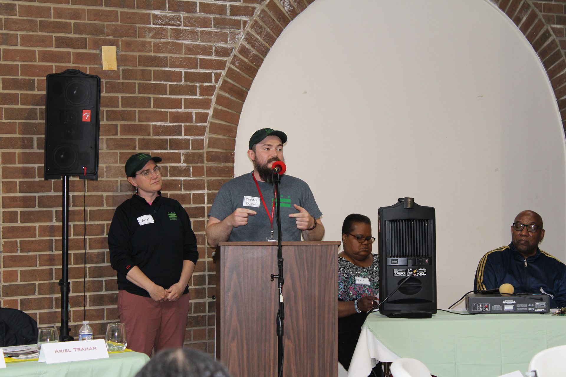 People at a podium, speaking at a conference. Brick wall backdrop. Other attendees seated.