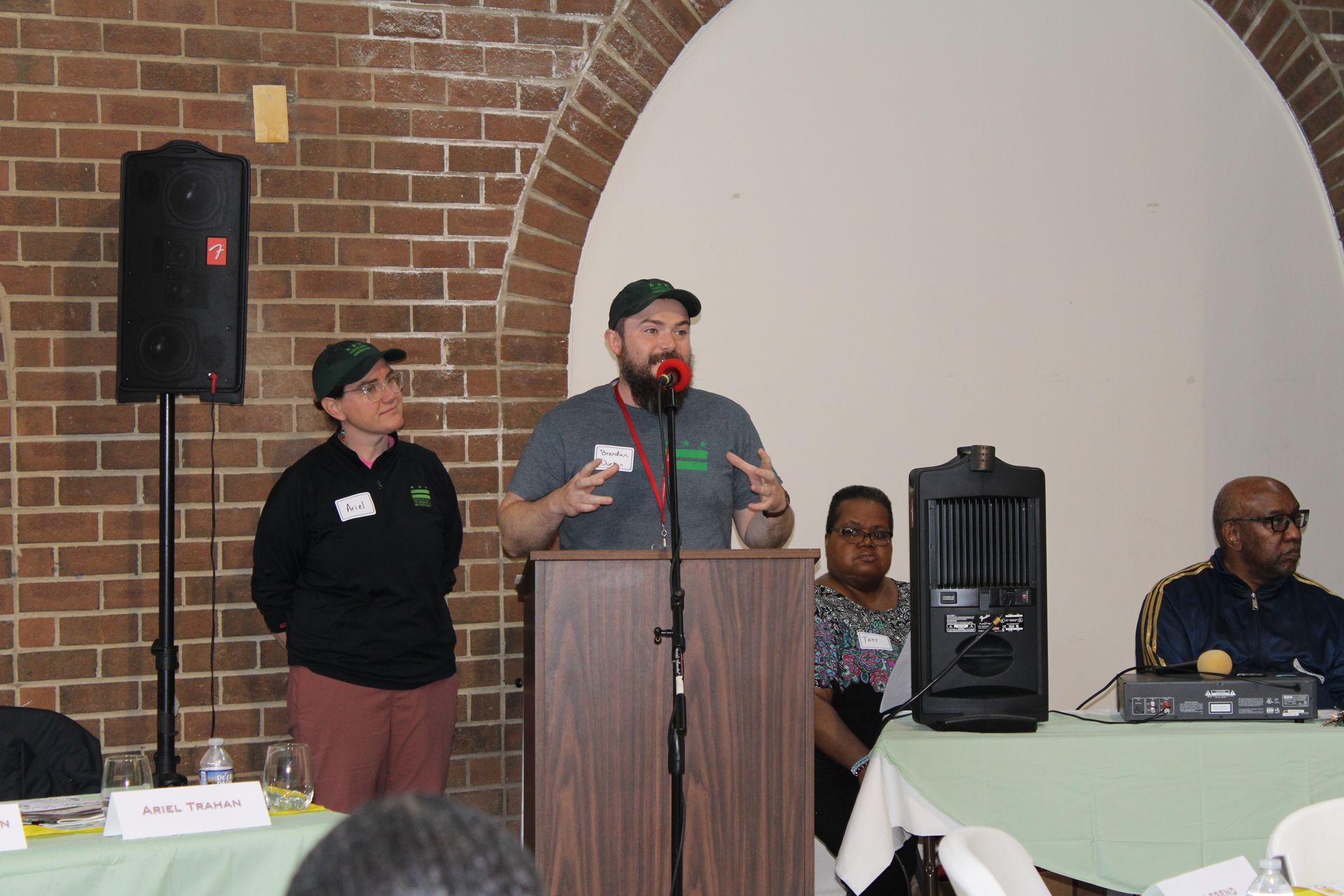 A man speaks at a podium; two others sit at a table, brick wall backdrop.