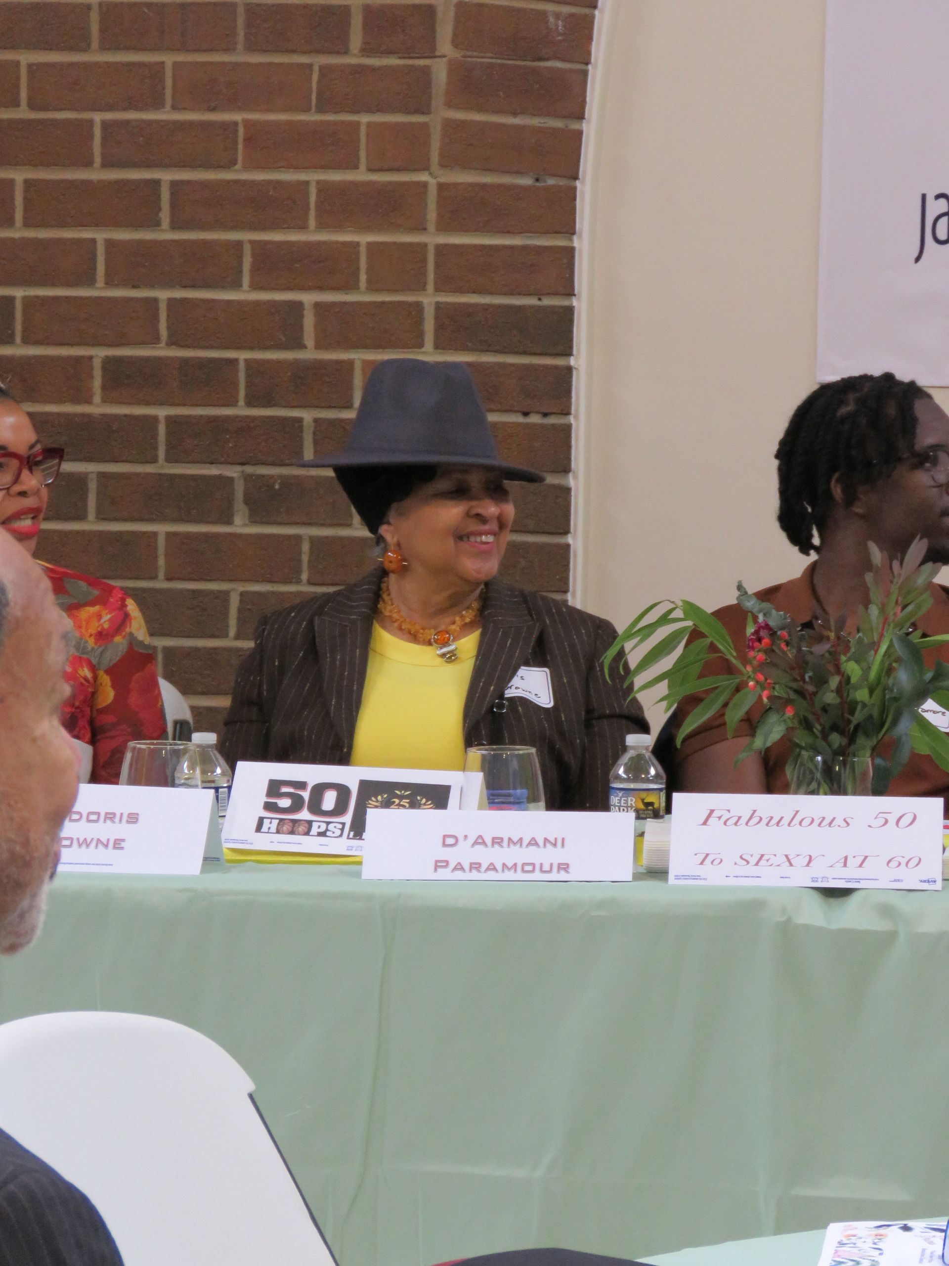 Three panelists seated at a table. Woman in center wears a hat and blazer. Brick wall background.