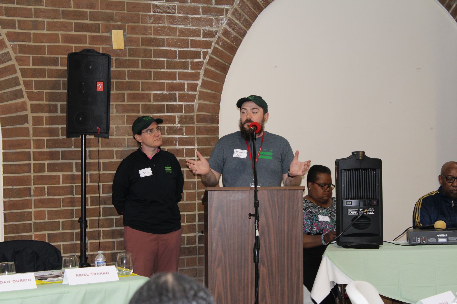 Two people at a podium in a brick-walled room with a speaker. One speaks, gesturing, the other stands beside.