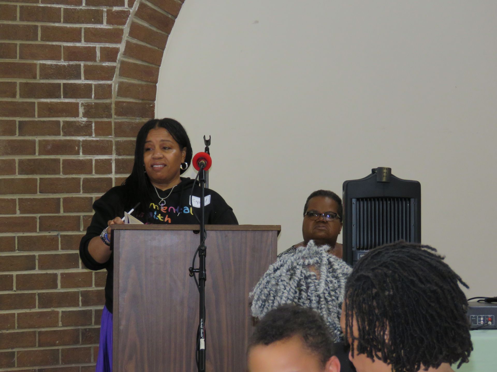 Woman speaking at a lectern, facing audience. Brick wall, white wall, and speakers are in the background.