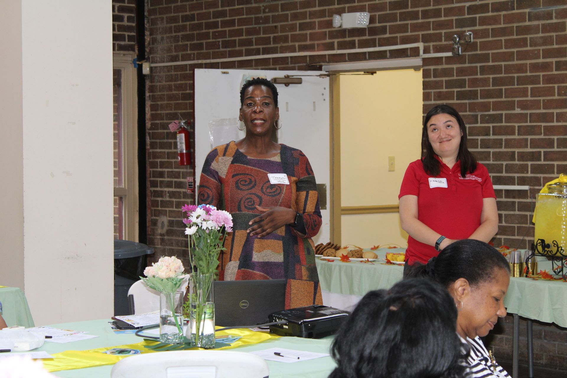 Two women at a table; one speaking, the other standing behind; refreshments and a flower vase are visible.