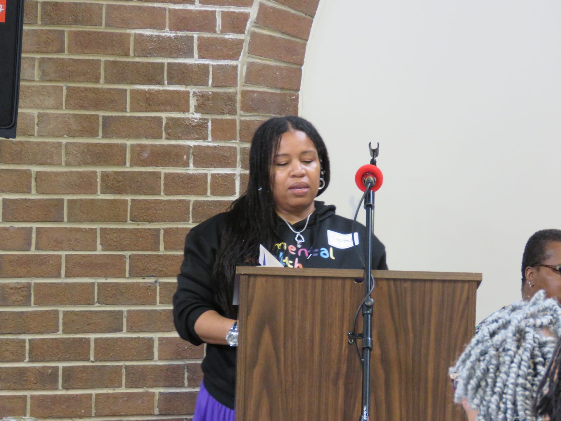 Woman speaking at a podium in front of a brick wall. She wears a black hoodie.