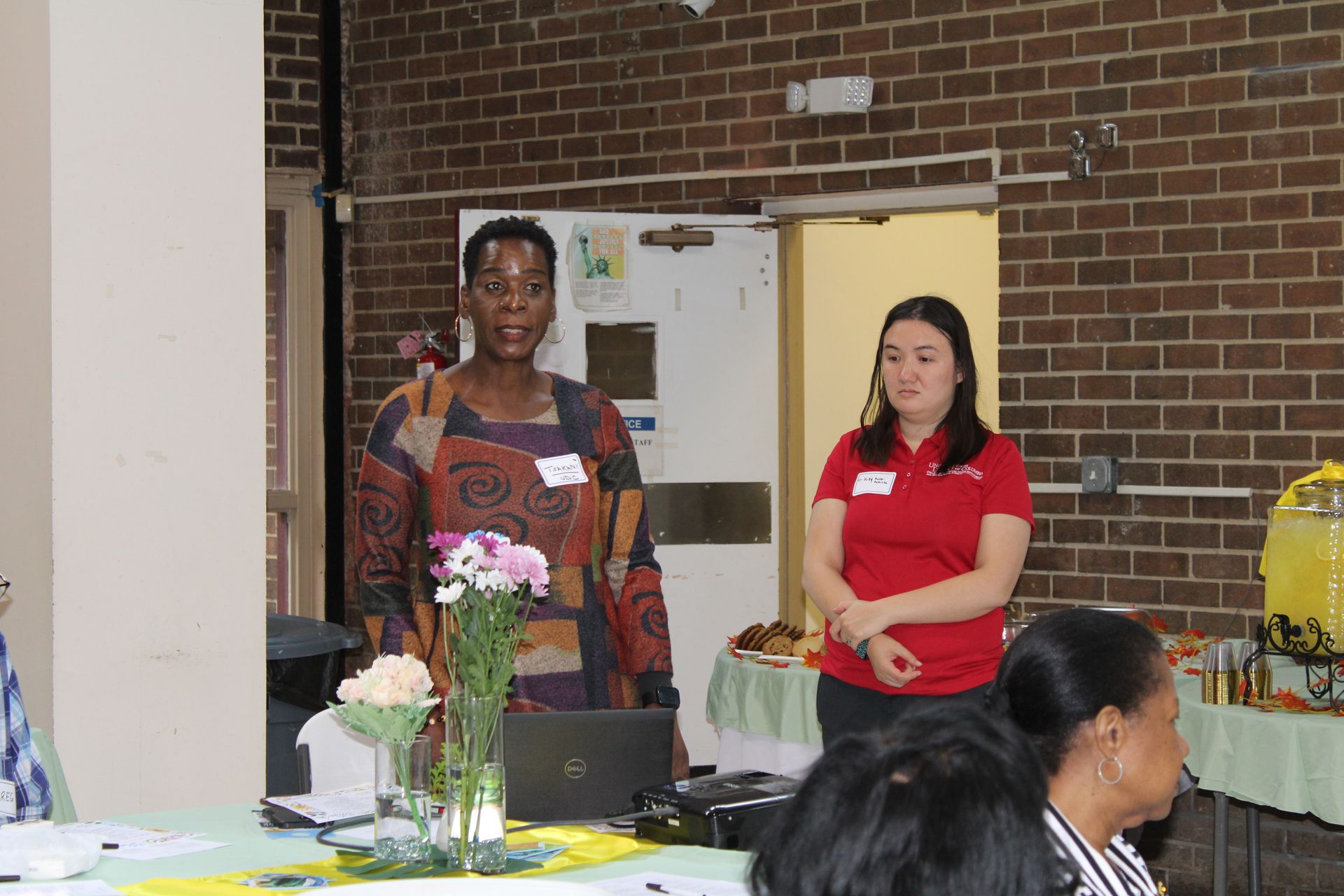 Two women at a table with flowers, one speaking, the other listening. Brick wall backdrop.