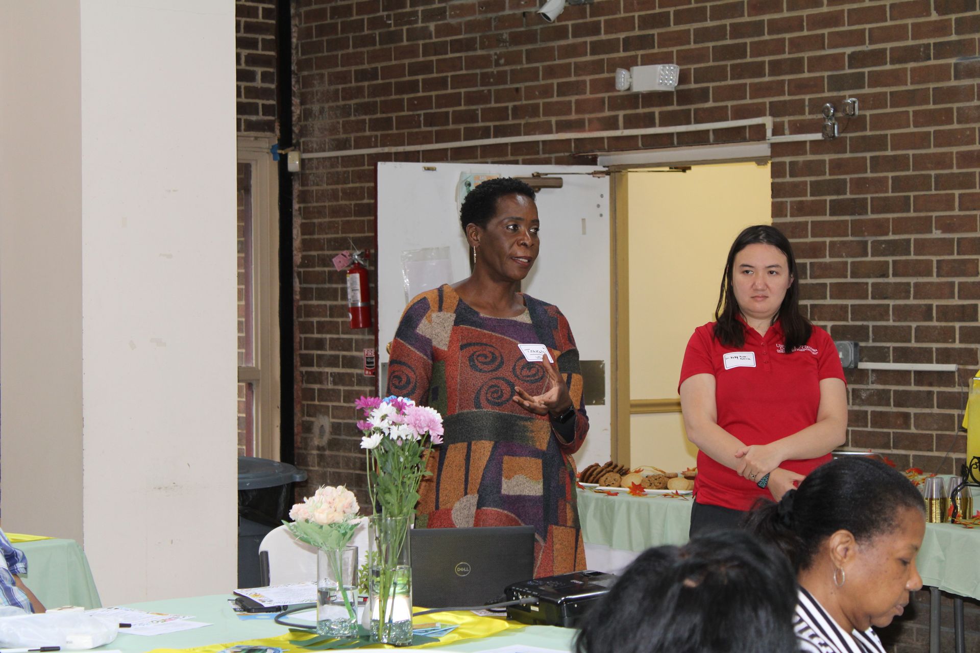 Woman speaking at podium, another woman stands beside. Indoor event with attendees.