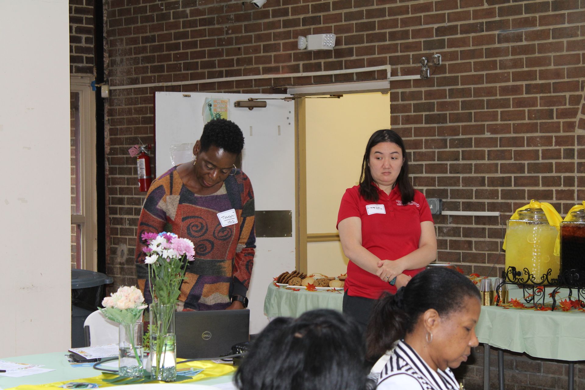 Two women at a presentation, a buffet spread visible. One woman speaks, the other stands by. Brick wall background.