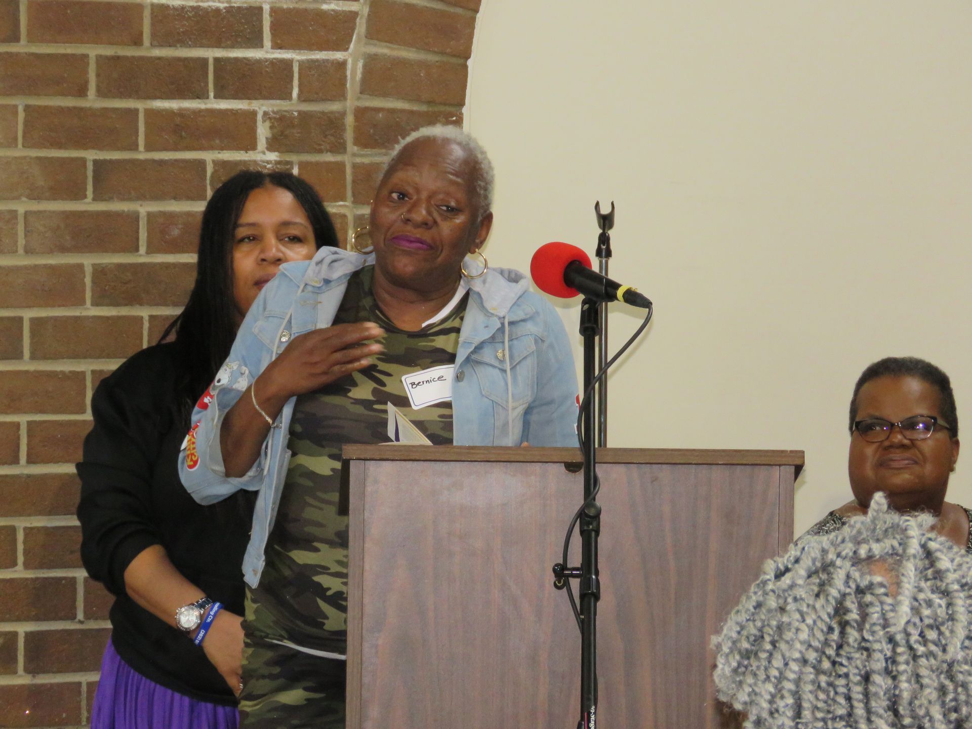 Woman speaking at a podium, wearing denim jacket and camouflage shirt. Two others stand nearby. Brick wall background.