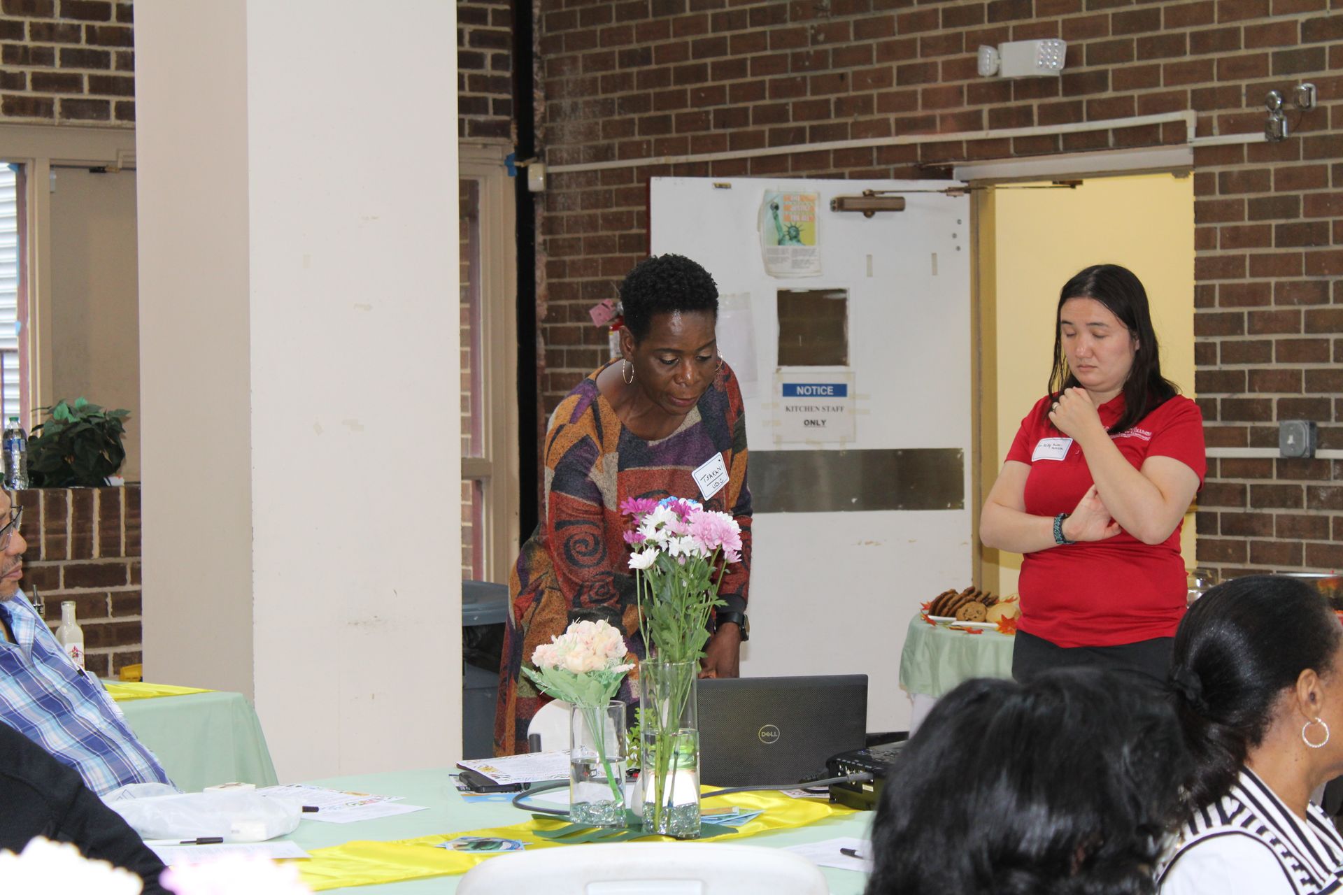 Two women at a presentation, one speaks near a laptop and flowers. The other watches with arms crossed. Brick wall backdrop.