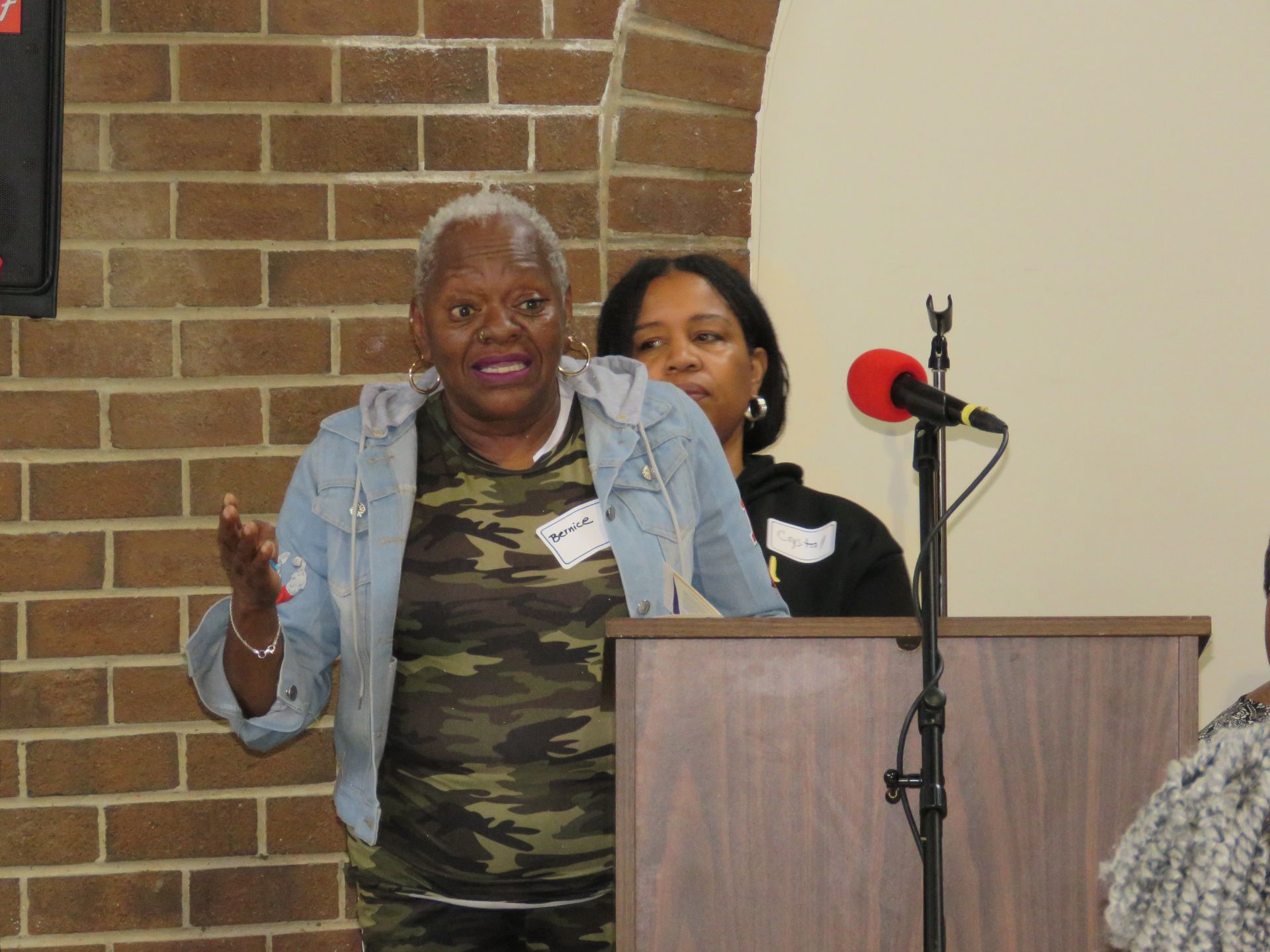 Woman speaking at a podium with microphone, another woman standing behind, brick wall in background.