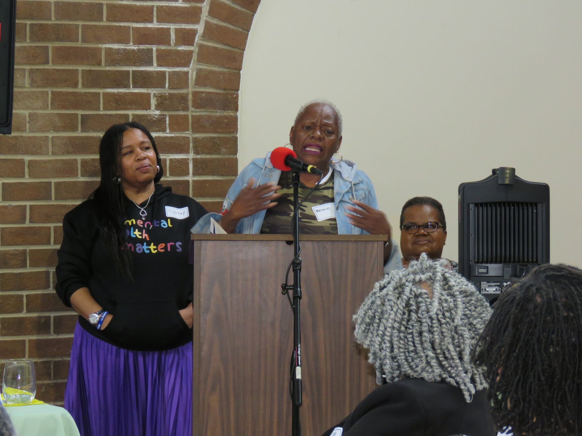 Three people at a podium with microphone. One speaks, another listens, and a third looks on. Brick wall in the background.