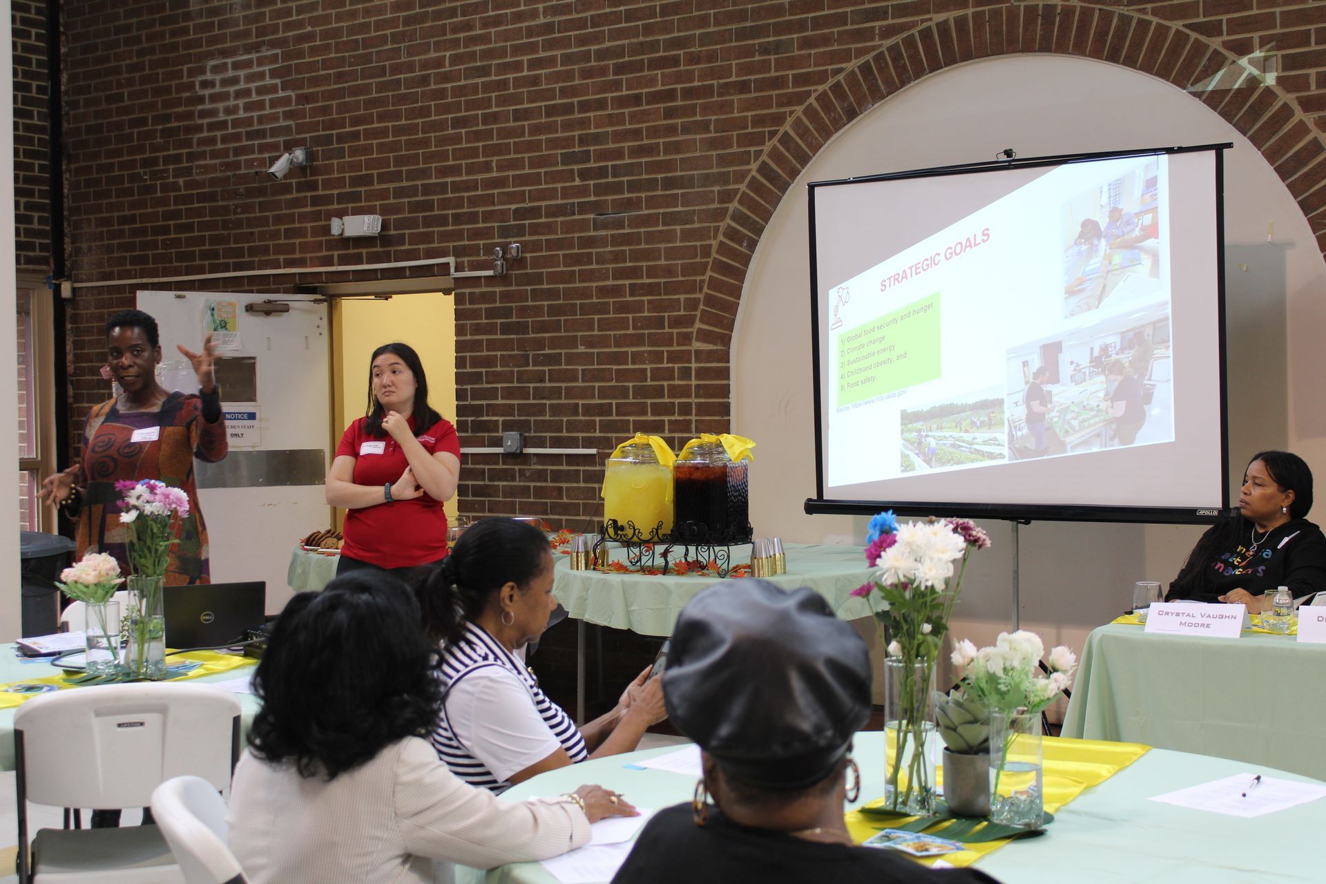 Presentation with three women speaking, slide show, and audience in a brick-walled room.