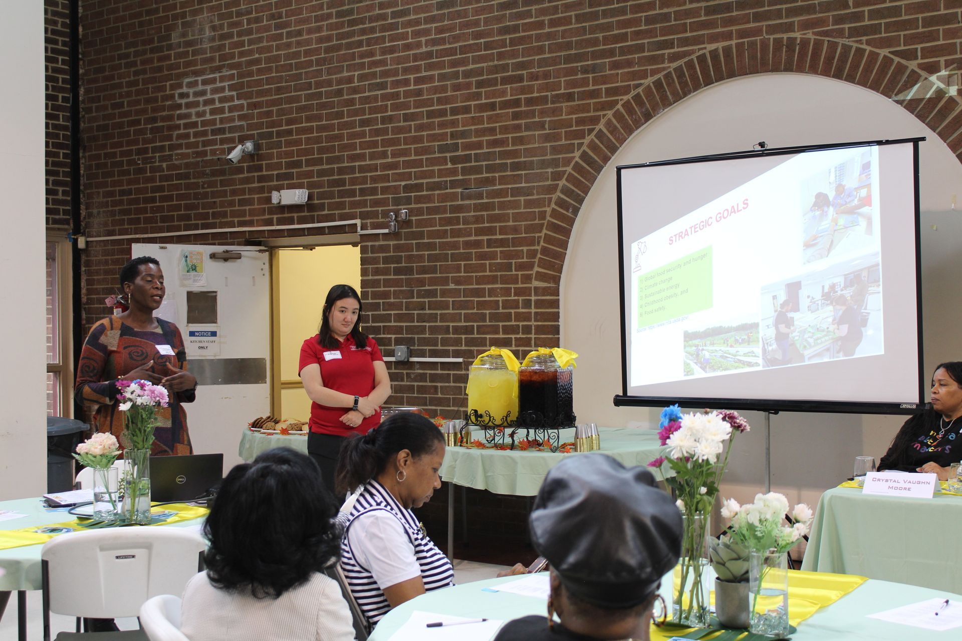 Two women presenting a slideshow in a room; audience seated at tables.