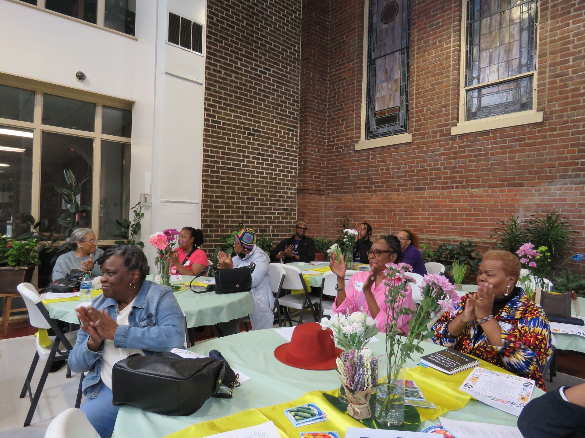 People clapping at tables set for an event, against a brick wall and window.